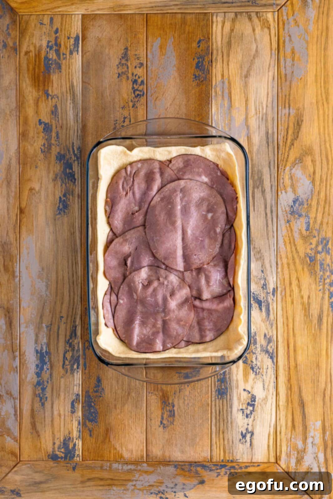 A baking dish displaying the layered ingredients: crescent dough, provolone cheese, and then a generous amount of roast beef.