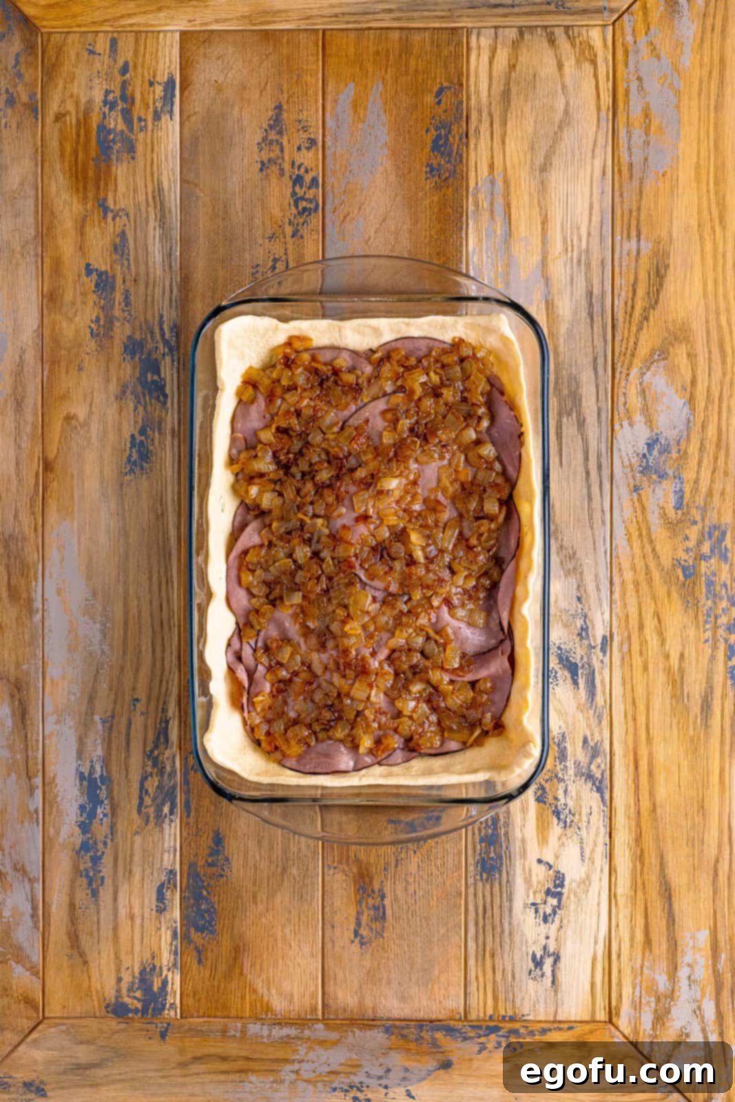 A baking dish layered with dough, cheese, roast beef, and a spread of golden, cooked onions on top.