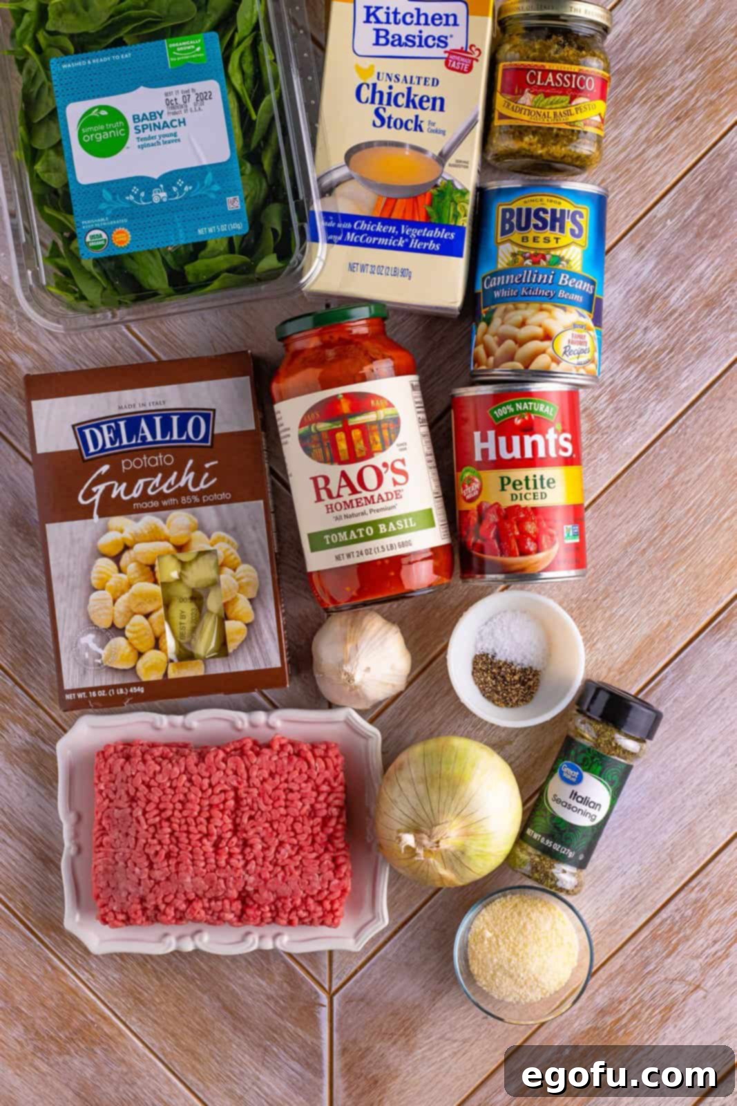 Various ingredients for Gnocchi Soup: Ground beef, sweet onion, garlic cloves, pesto, Italian seasoning, kosher salt, black pepper, chicken stock, pasta sauce, cannelini beans, diced tomatoes, gnocchi, baby spinach, and parmesan cheese arranged on a wooden board.