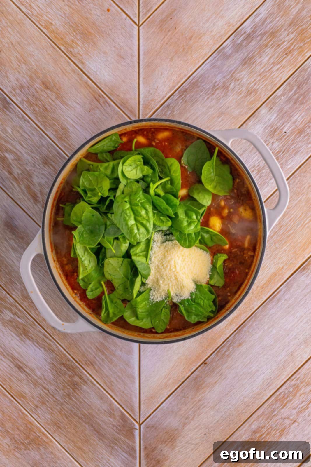 Spinach and grated Parmesan cheese being stirred into a pot of rich tomato-based soup.
