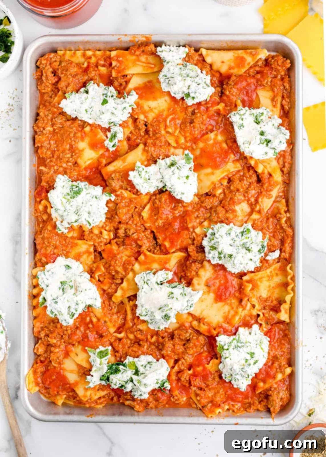 A sheet pan of lasagna base with dollops of white ricotta cheese mixture artfully placed on top.