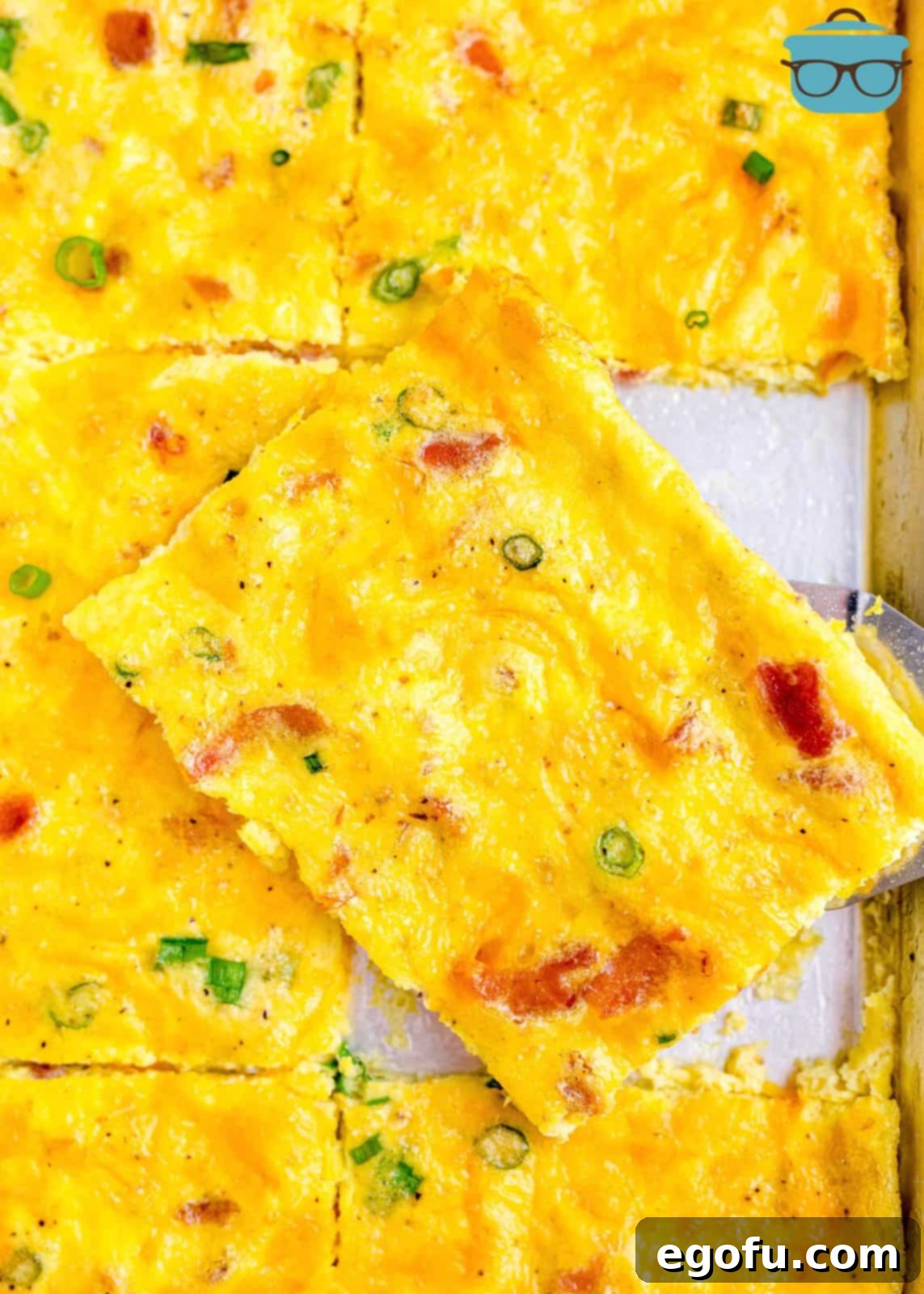 Baked Sheet Pan Eggs 2 A perfectly baked slice of Sheet Pan Eggs, slightly tilted to showcase its fluffy interior and rich blend of ham, bacon, and cheese, resting on a bed of more eggs on the baking pan.
