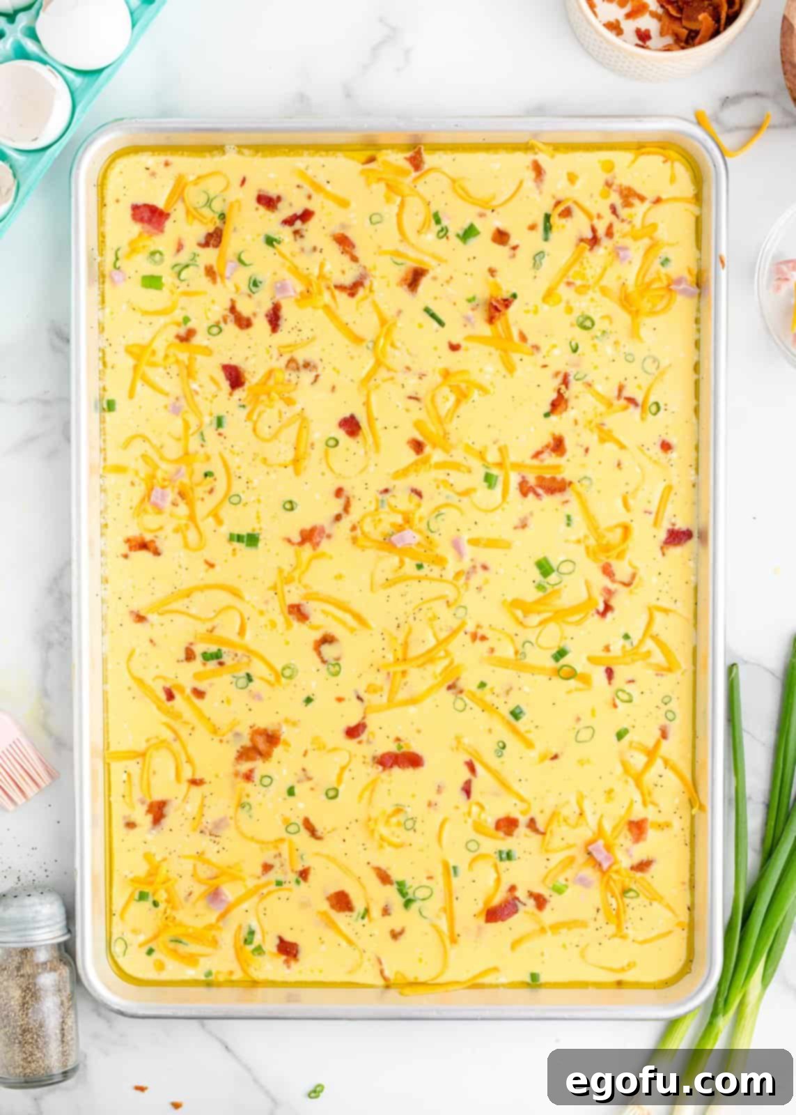 Baked Sheet Pan Eggs 7 A sheet pan with the liquid egg mixture generously topped with colorful cubed ham, crumbled bacon, vibrant diced green onions, and savory shredded cheese, perfectly arranged before baking.