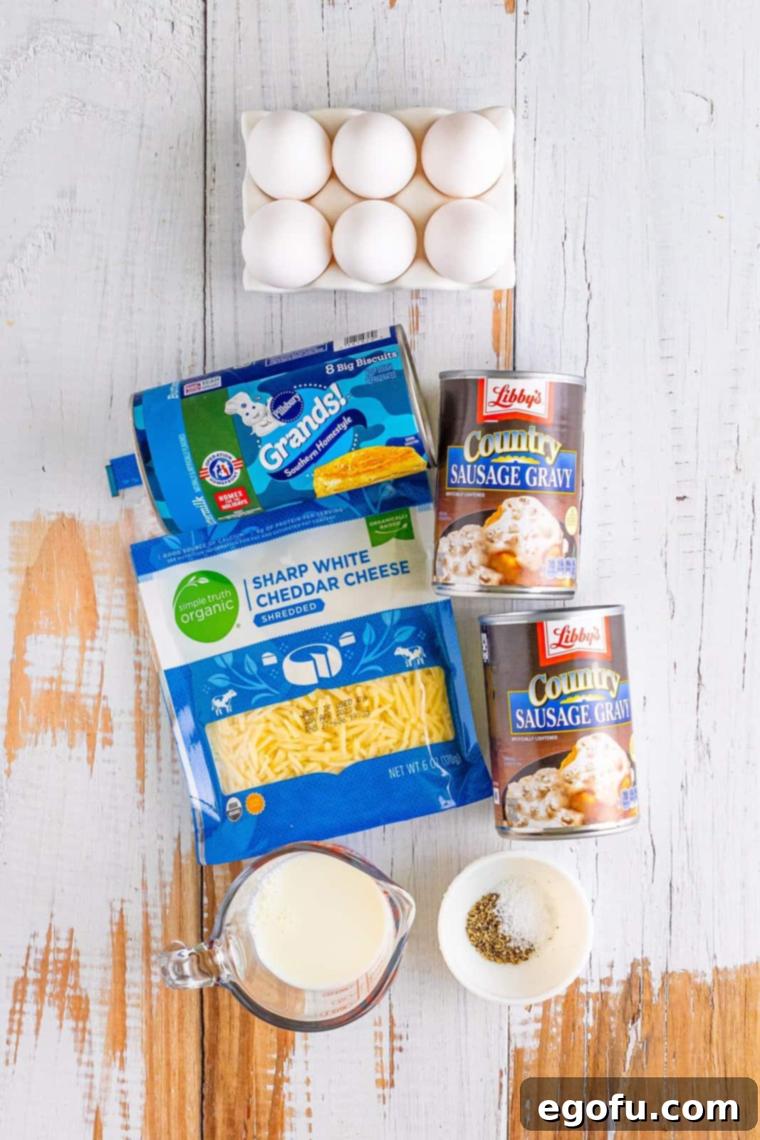 Biscuits, eggs, white cheddar cheese, milk, salt, pepper, and cans of sausage gravy laid out as ingredients.