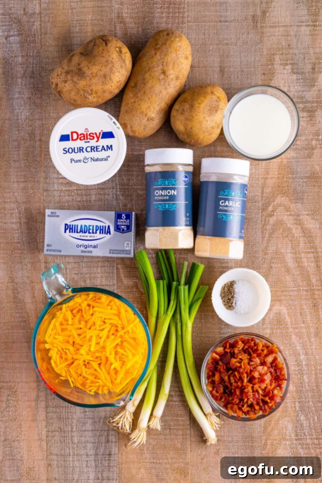 A vibrant display of all the ingredients needed for the Twice Baked Potato Casserole: whole potatoes, containers of sour cream and cream cheese, bundles of green onions, strips of bacon, garlic powder, shredded cheddar cheese, milk, onion powder, kosher salt, and black pepper.