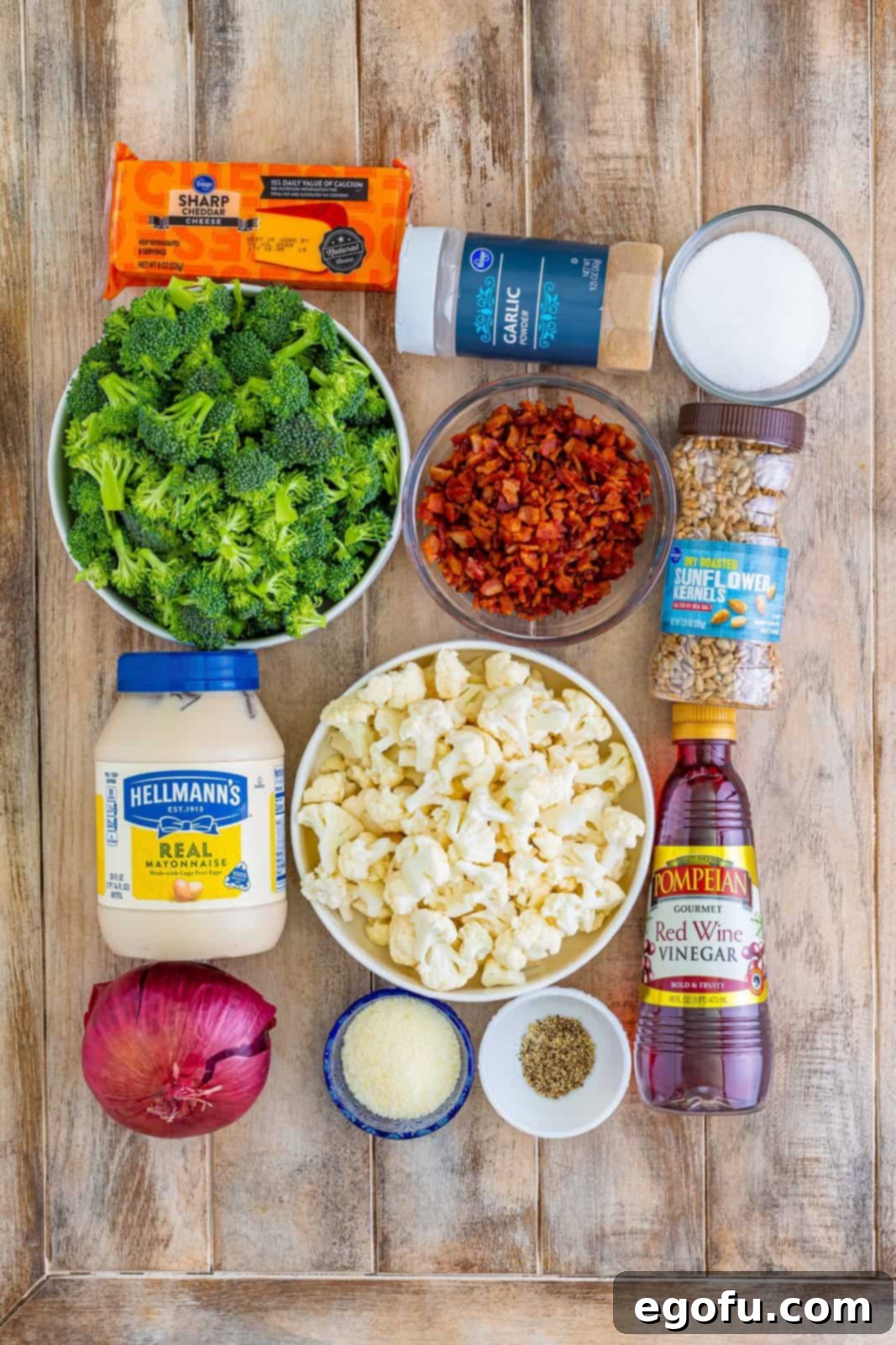 A flat lay of all the fresh ingredients for the broccoli cauliflower salad: mayonnaise, sugar, broccoli, cauliflower, red onion, parmesan cheese, red wine vinegar, garlic powder, black pepper, sunflower seeds, bacon, and sharp cheddar cheese cubes.