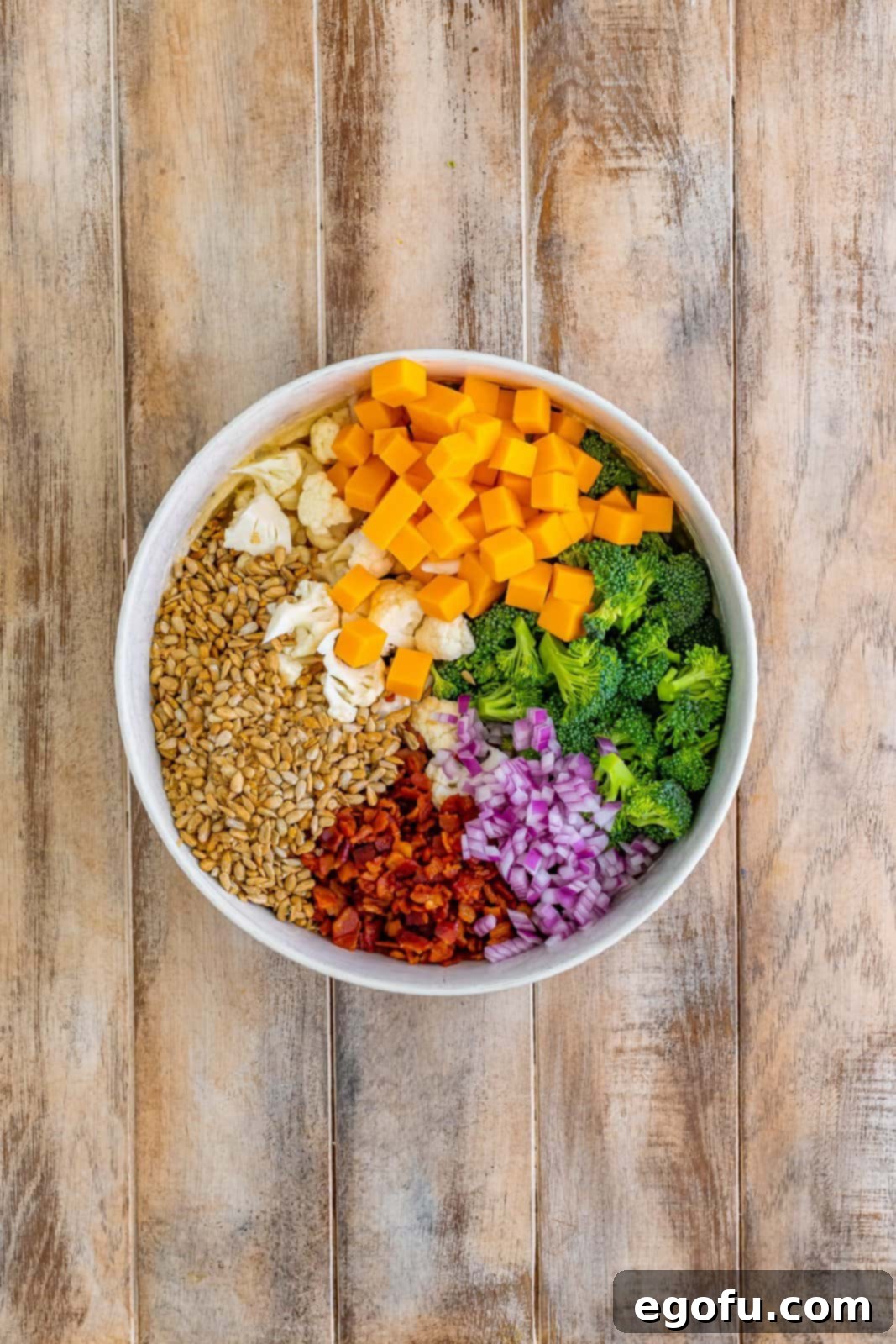 A large mixing bowl containing the dressing, with fresh broccoli, cauliflower, cooked bacon, cheddar cheese cubes, sunflower seeds, and red onion added on top.