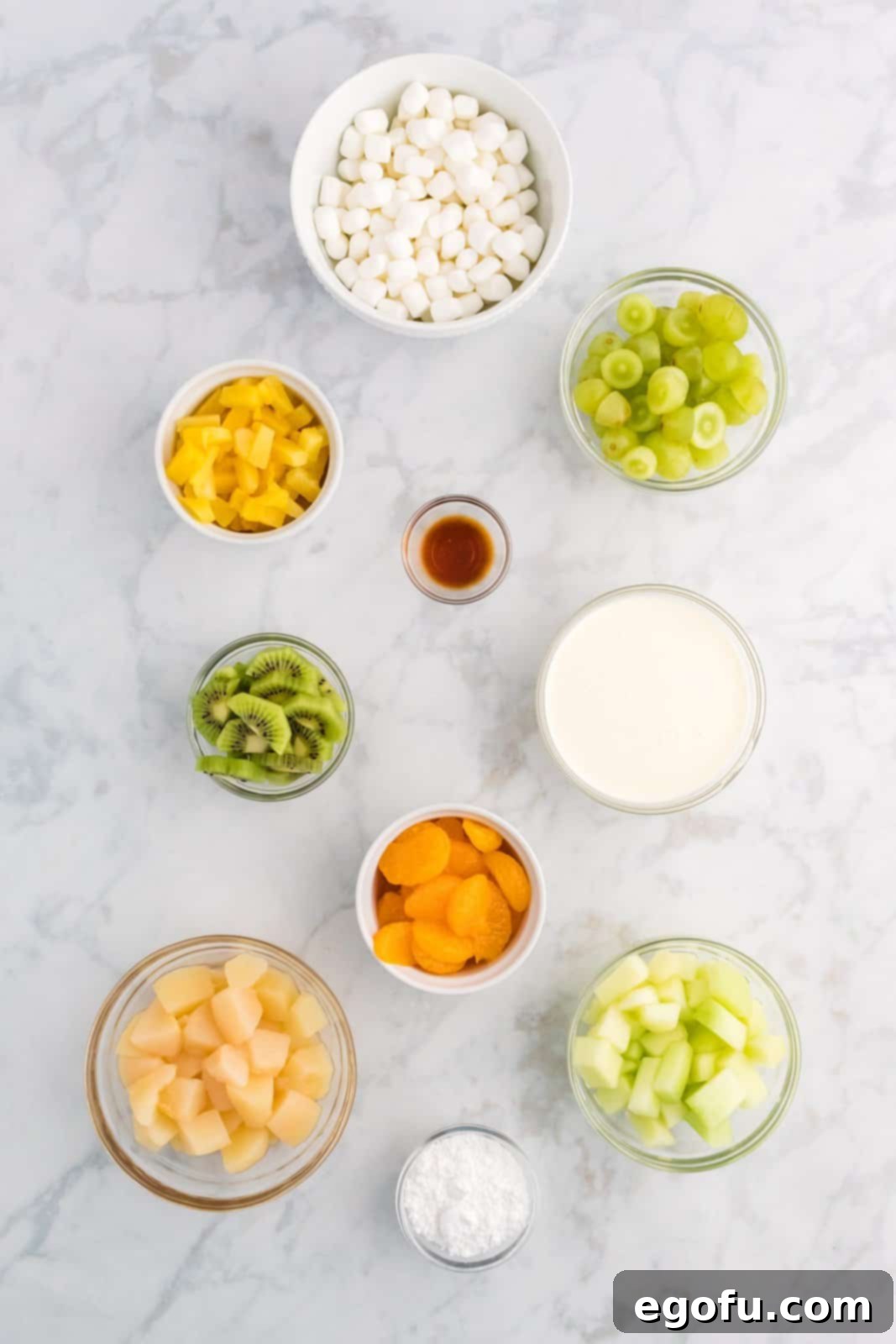 Emerald and Golden Ambrosia Delight 4 An array of ingredients for Green and Gold Ambrosia Salad, including heavy whipping cream, powdered sugar, vanilla extract, mini marshmallows, sliced pears, mandarin oranges, kiwis, green grapes, honeydew melon, and pineapple tidbits.