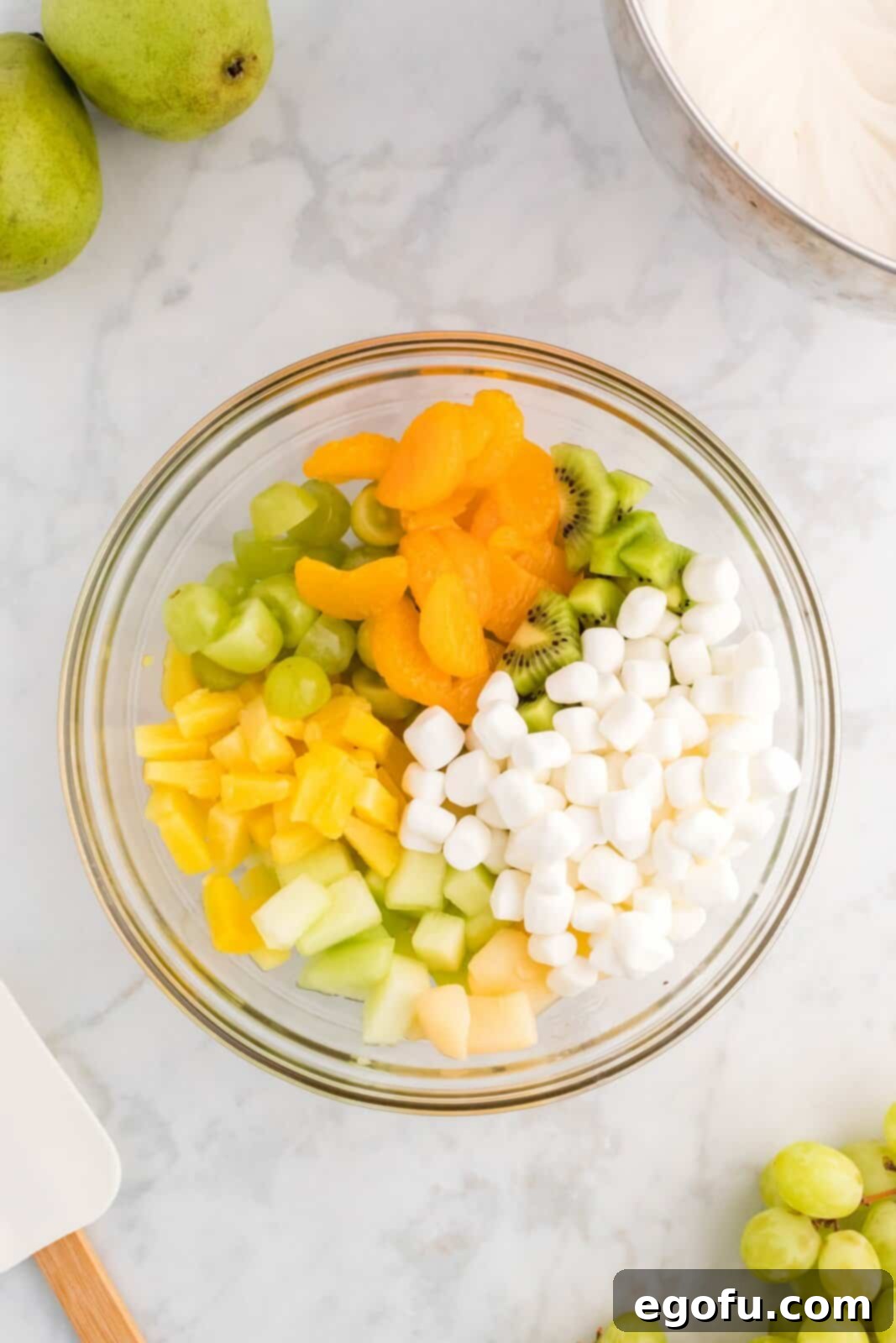 Emerald and Golden Ambrosia Delight 6 A large mixing bowl containing a colorful assortment of green and gold fruits along with mini marshmallows, ready for the creamy dressing.