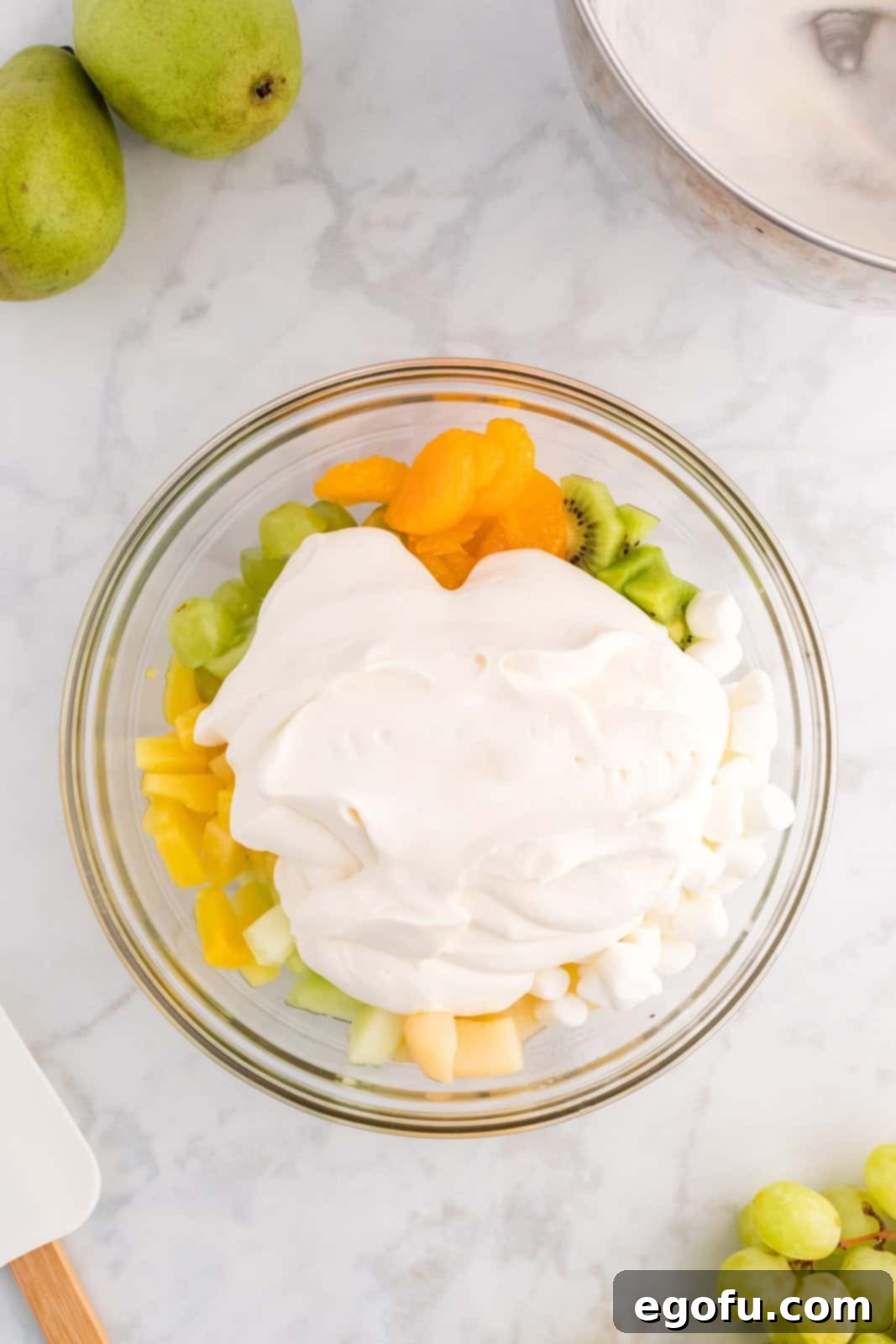 Emerald and Golden Ambrosia Delight 7 A generous dollop of homemade whipped cream placed on top of the fruit and marshmallow mixture in a large mixing bowl.