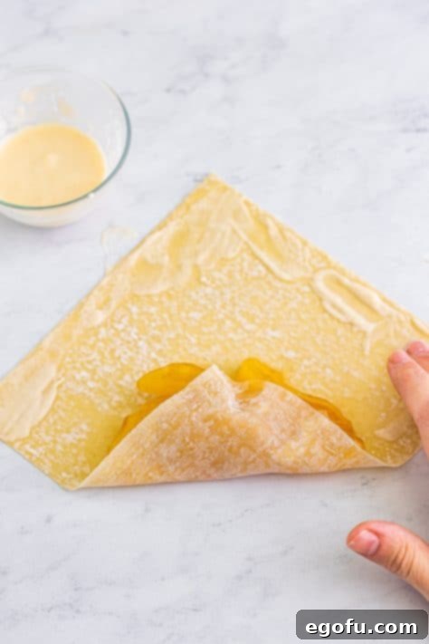 A moistened egg roll wrapper with apple pie filling that is being rolled up.