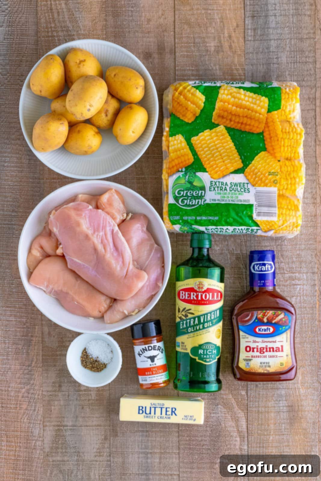 One-Pan BBQ Chicken Dinner 4 Individual ingredients laid out: chicken breasts, a bottle of BBQ sauce, Yukon Gold potatoes, mini corn on the cobs, a bowl of salt and pepper, BBQ seasoning, and a bottle of olive oil.