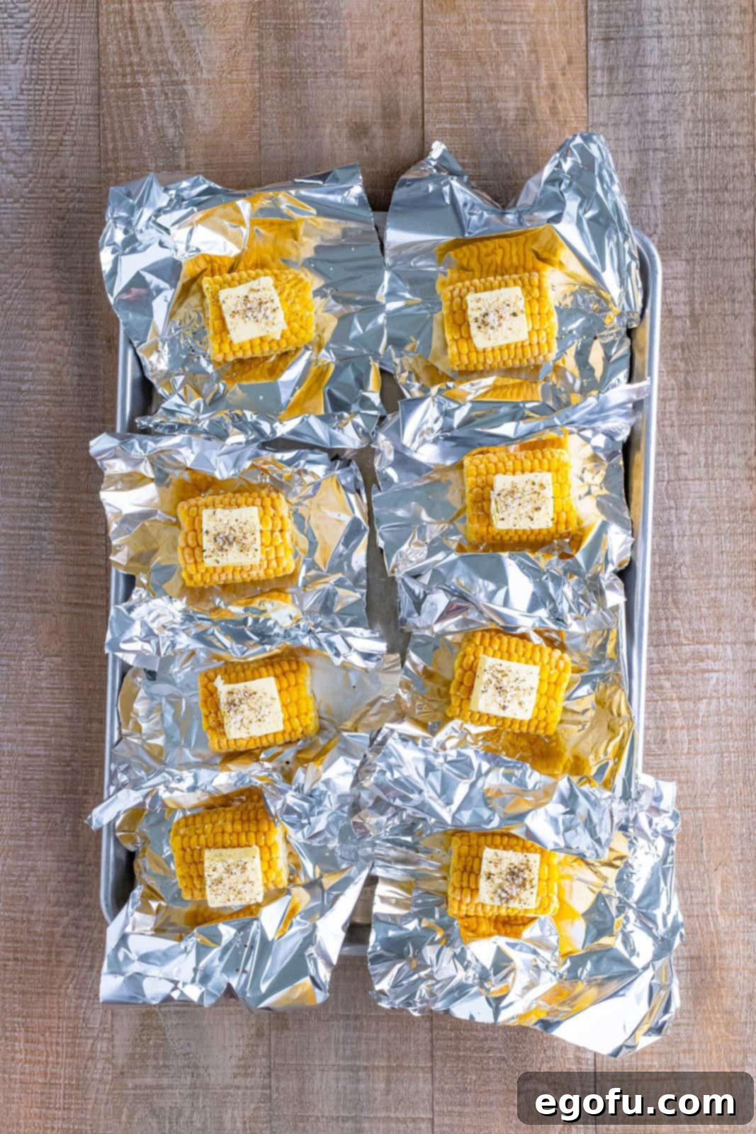 One-Pan BBQ Chicken Dinner 5 Pieces of aluminum foil with mini cobs of corn, each topped with a pat of butter and seasoning, ready to be wrapped.