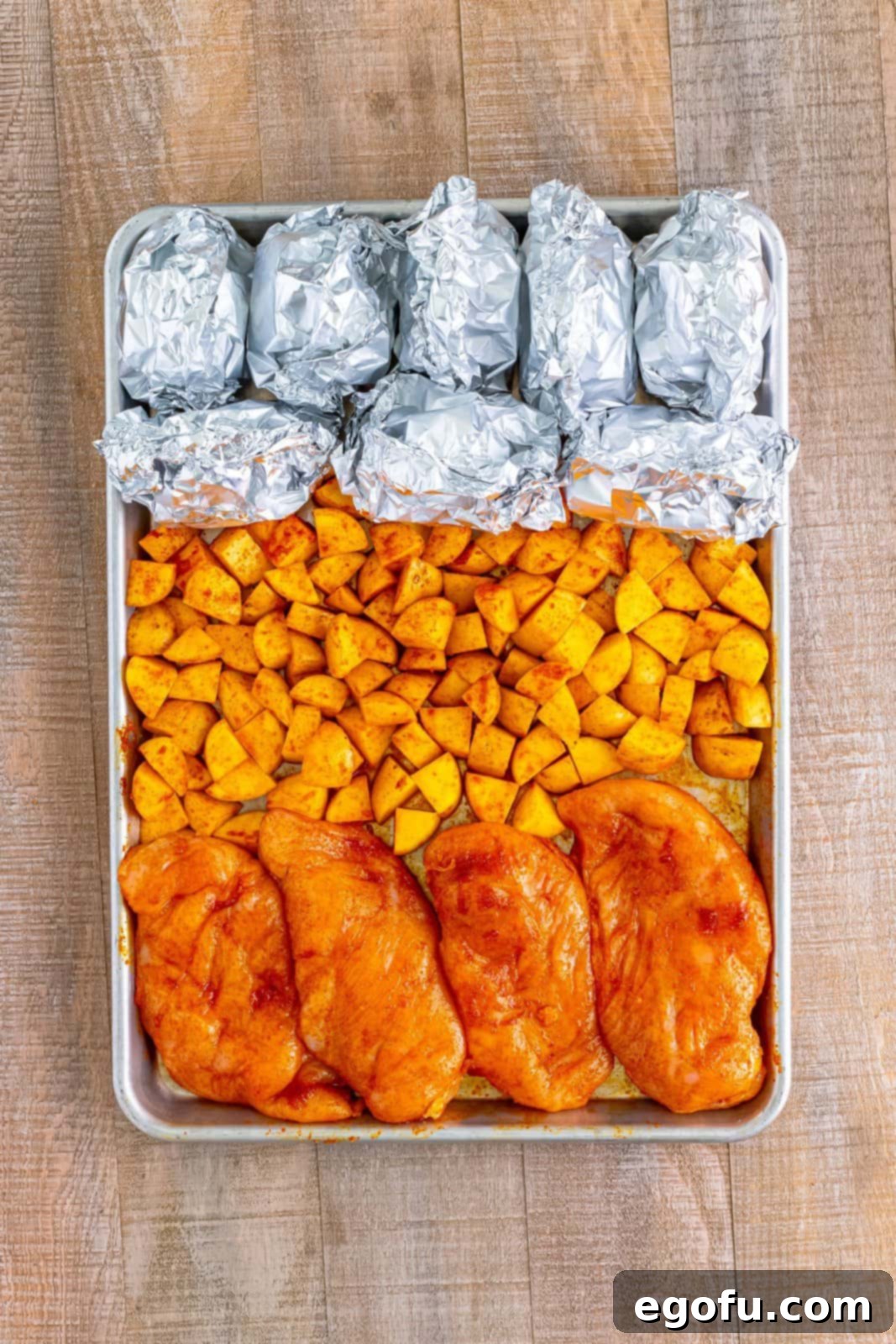 One-Pan BBQ Chicken Dinner 8 A sheet pan with seasoned chicken breasts, diced potatoes, and aluminum foil-covered corn on the cob, ready for baking.