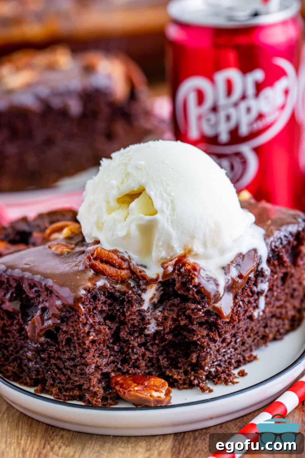A slice of Dr Pepper cake on a small round white plate with a scoop of vanilla ice cream slightly melted on top of the cake, with a small can of Dr Pepper soda in the background.