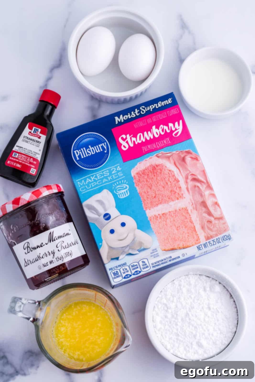 Strawberry Swirl Brownie Bliss 4 A flat lay of all the essential ingredients for making Strawberry Brownies: strawberry cake mix, a bottle of strawberry extract, fresh eggs, a jar of strawberry preserves, a bowl of melted butter, a glass of milk, and a bowl of powdered sugar.