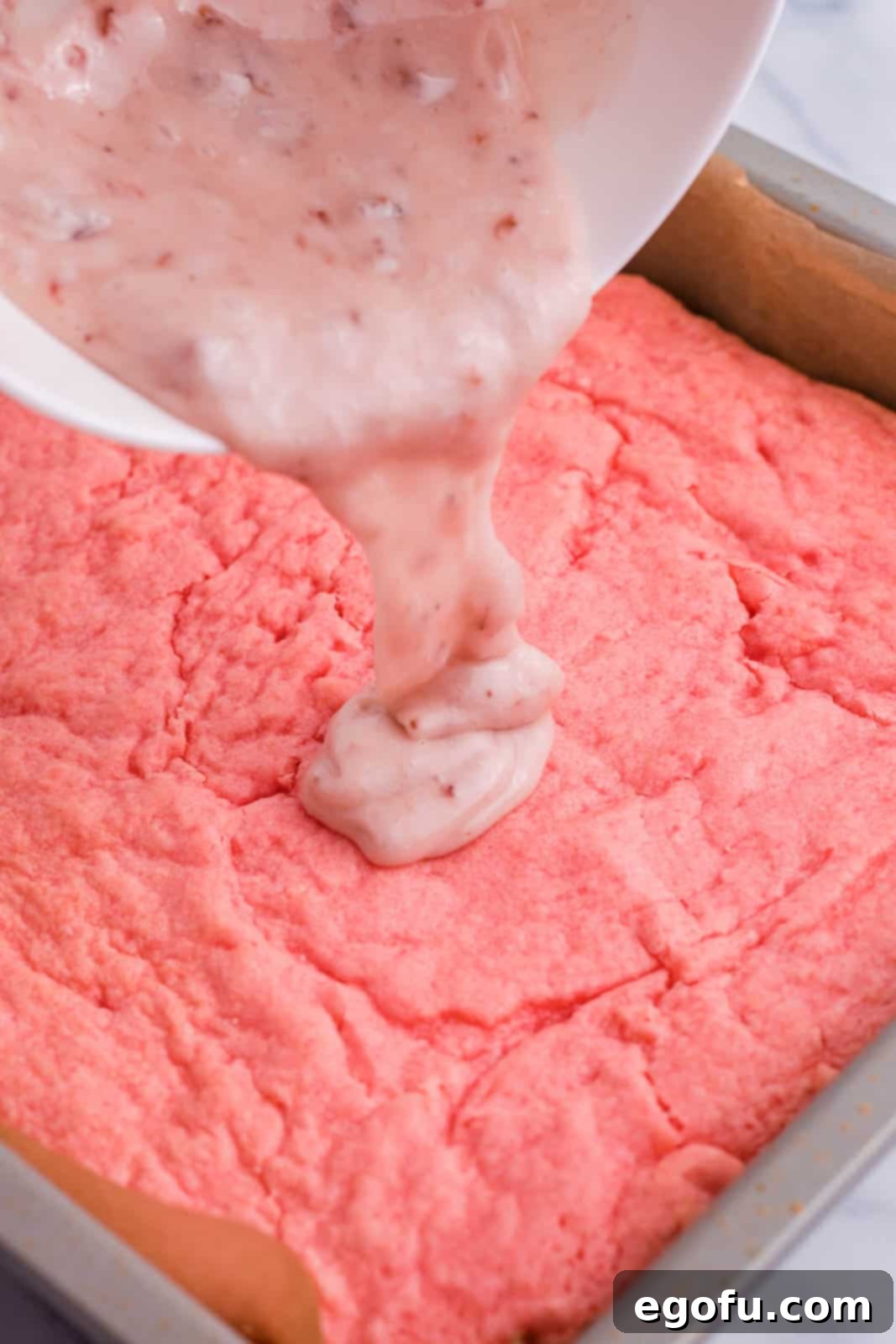 Strawberry Swirl Brownie Bliss 8 A sweet, pink strawberry glaze being poured generously over the top of cooled strawberry brownies in a pan, ready to be spread evenly.