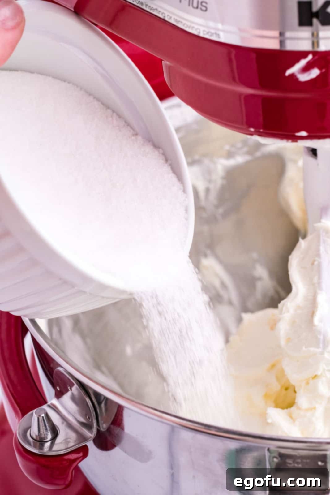 A cup pouring in sugar into a mixing bowl with creamed cream cheese.
