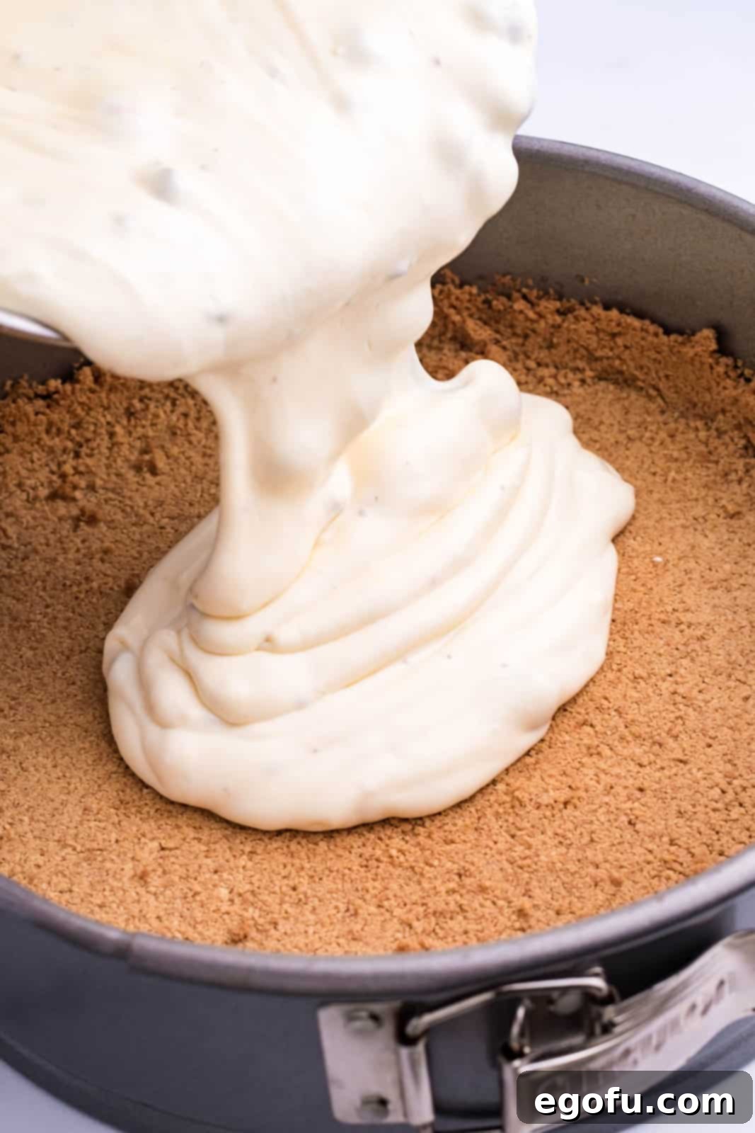 Cheesecake filling being poured into a graham cracker crumb crust in a pan.