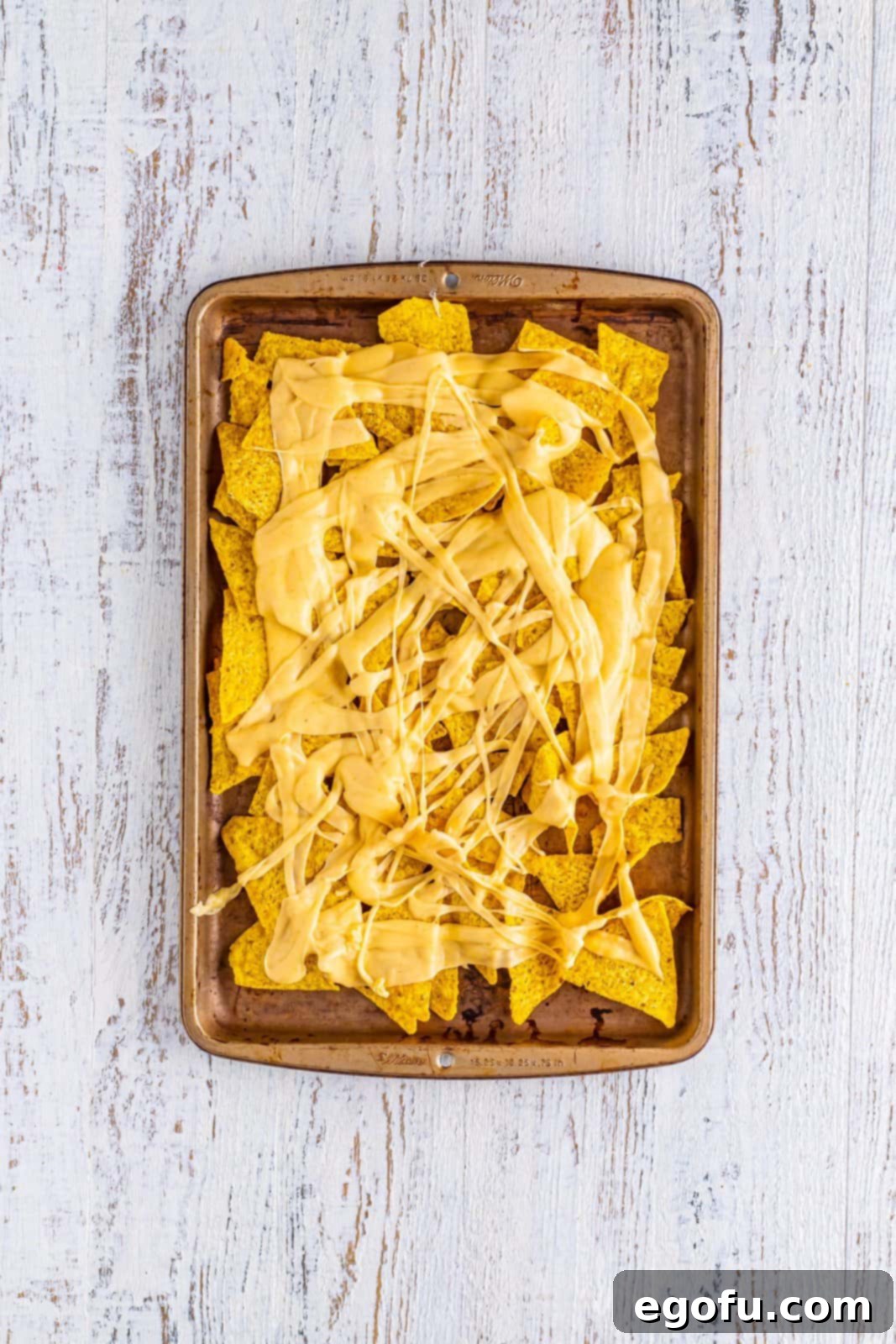 A layer of tortilla chips on a sheet tray, glistening with a drizzle of melted cheese sauce.
