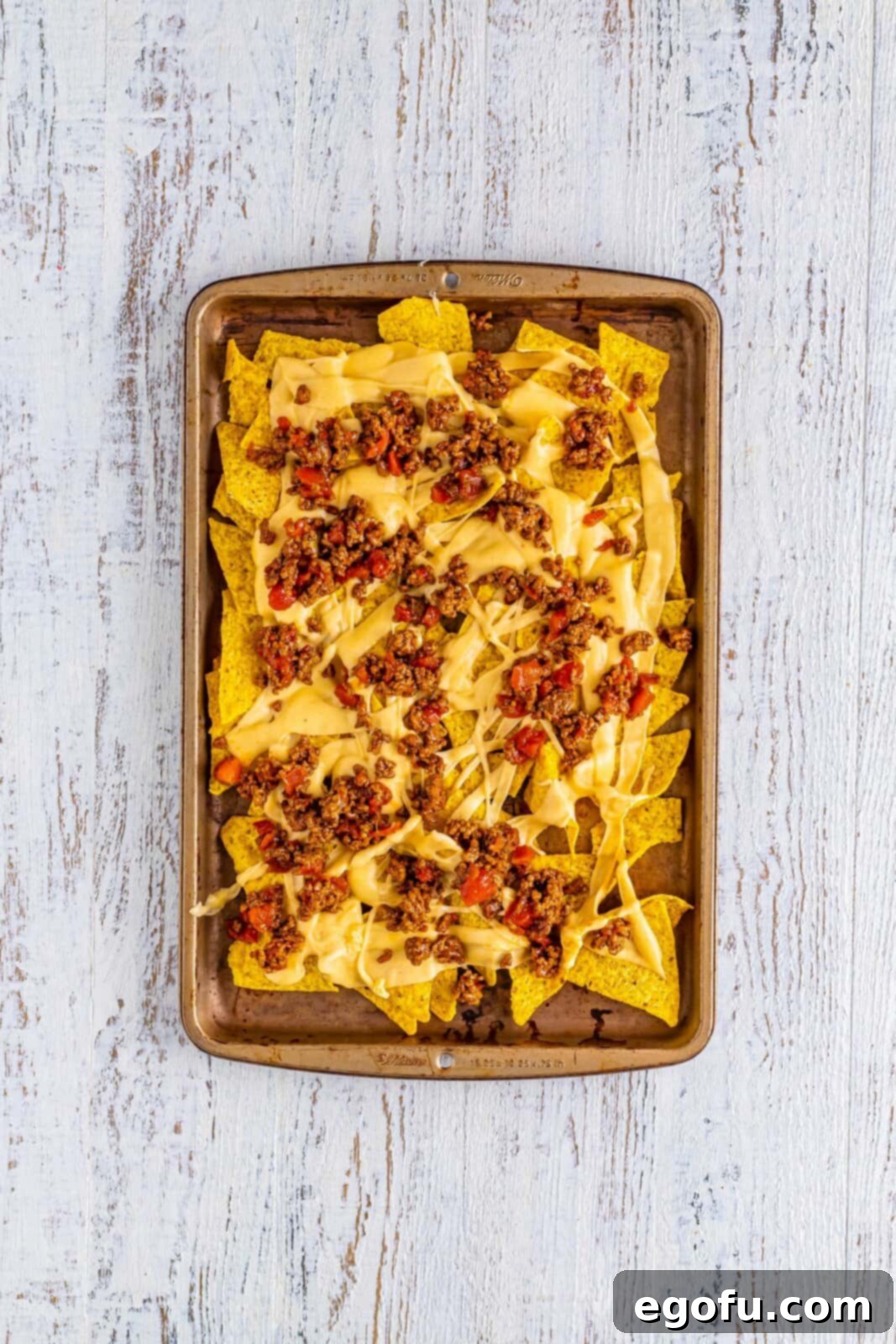 The first layer of nachos on a sheet tray, showing tortilla chips, cheese sauce, and a topping of seasoned ground beef with Rotel.