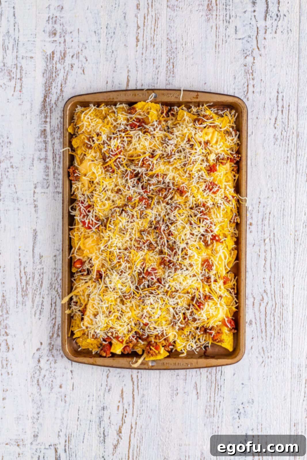 Fully layered nachos on a sheet tray, showing multiple levels of chips, meat, and cheese, ready for baking.