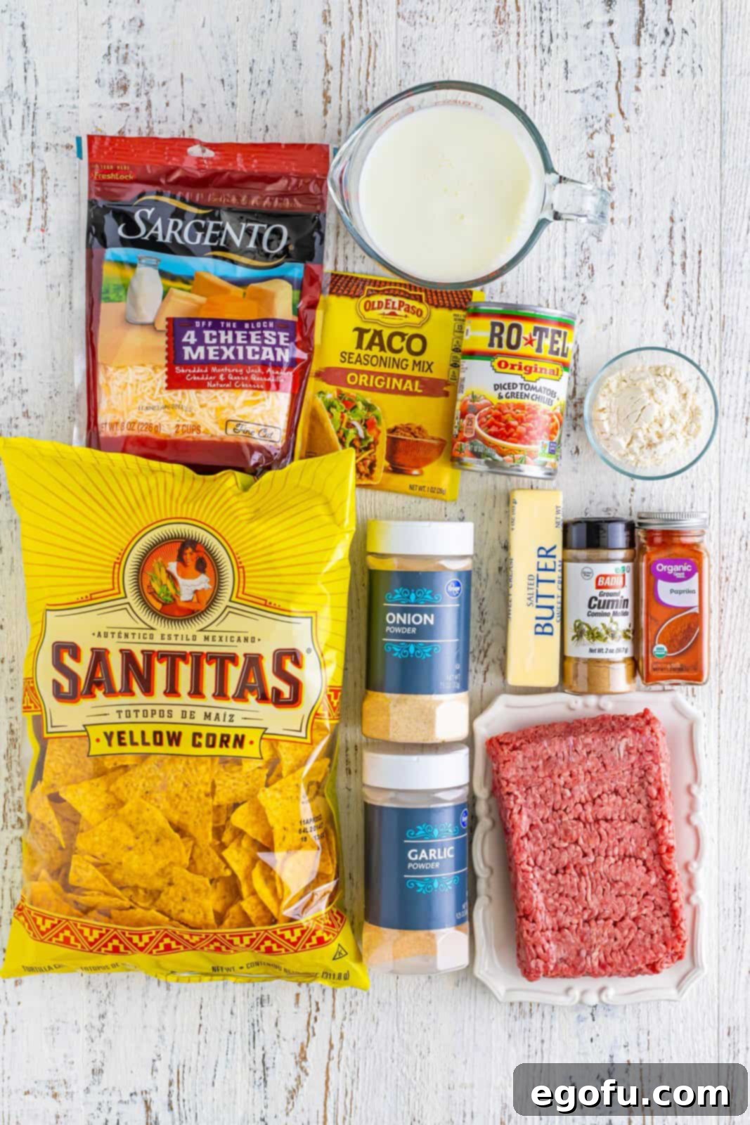 A flat lay photograph displaying all the fresh ingredients needed for sheet pan nachos: ground beef, taco seasoning, Rotel, salted butter, all-purpose flour, garlic powder, onion powder, ground cumin, sweet paprika, milk, shredded Mexican blend cheese, tortilla chips, diced tomatoes, sour cream, sliced black olives, and fresh chopped cilantro.