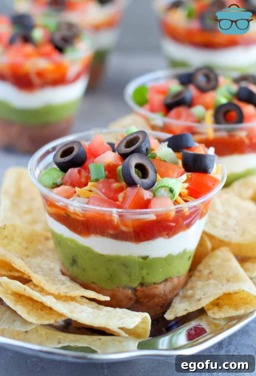 A plastic cup with a layered dip and chips around it on a plate. 