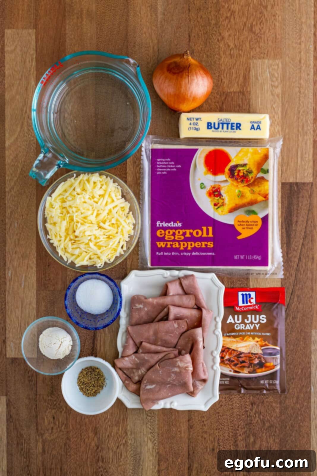 A flat lay of all ingredients for French Dip Egg Rolls: butter, onion, sugar, pepper, water, flour, egg roll wrappers, roast beef slices, provolone cheese, and au jus gravy mix.