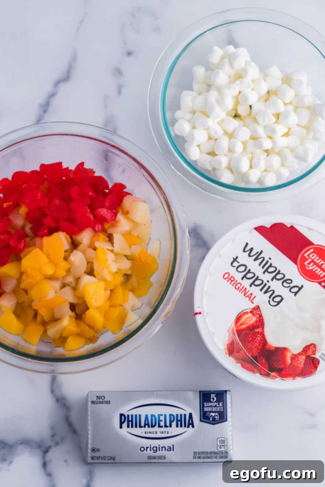 Individual ingredients for frozen fruit salad laid out: softened cream cheese, thawed whipped topping, mini marshmallows, canned fruit cocktail, and maraschino cherries.