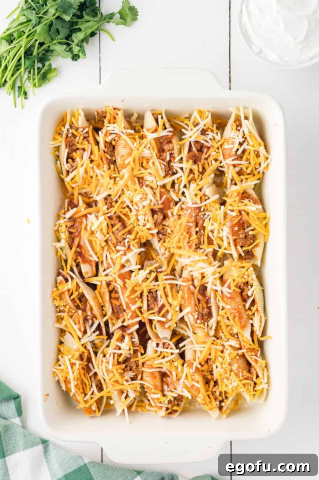 Baking dish with taco stuffed shells, now fully prepared with enchilada sauce and a thick layer of shredded cheese, ready for the oven.