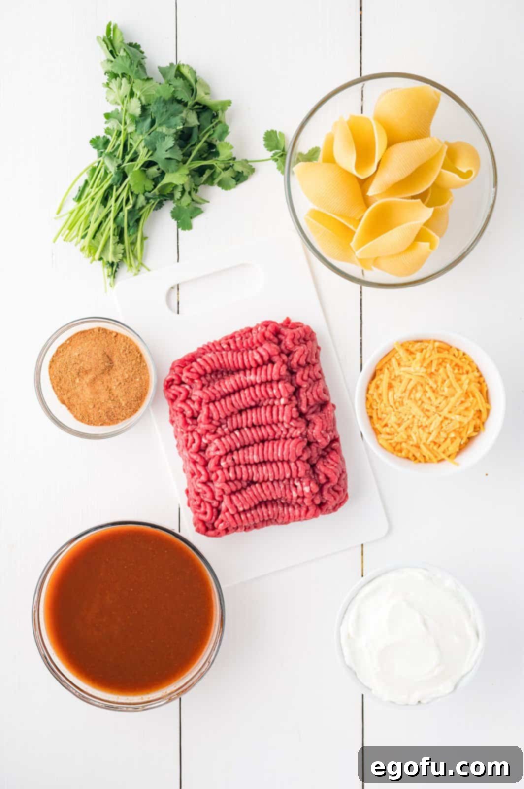 A flat lay displaying all the essential ingredients for Taco Stuffed Shells: ground beef, fresh cilantro, a can of enchilada sauce, shredded cheese, taco seasoning, sour cream, and dry jumbo pasta shells.