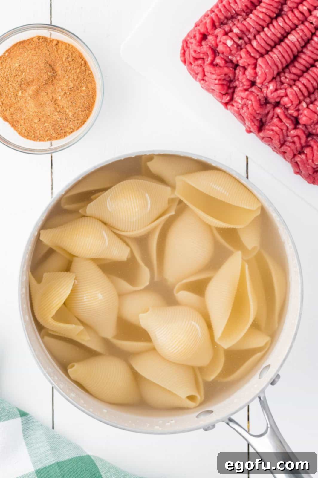 Skillet with jumbo pasta shells cooking, while raw ground beef and taco seasoning are placed nearby, representing the preparation phase.