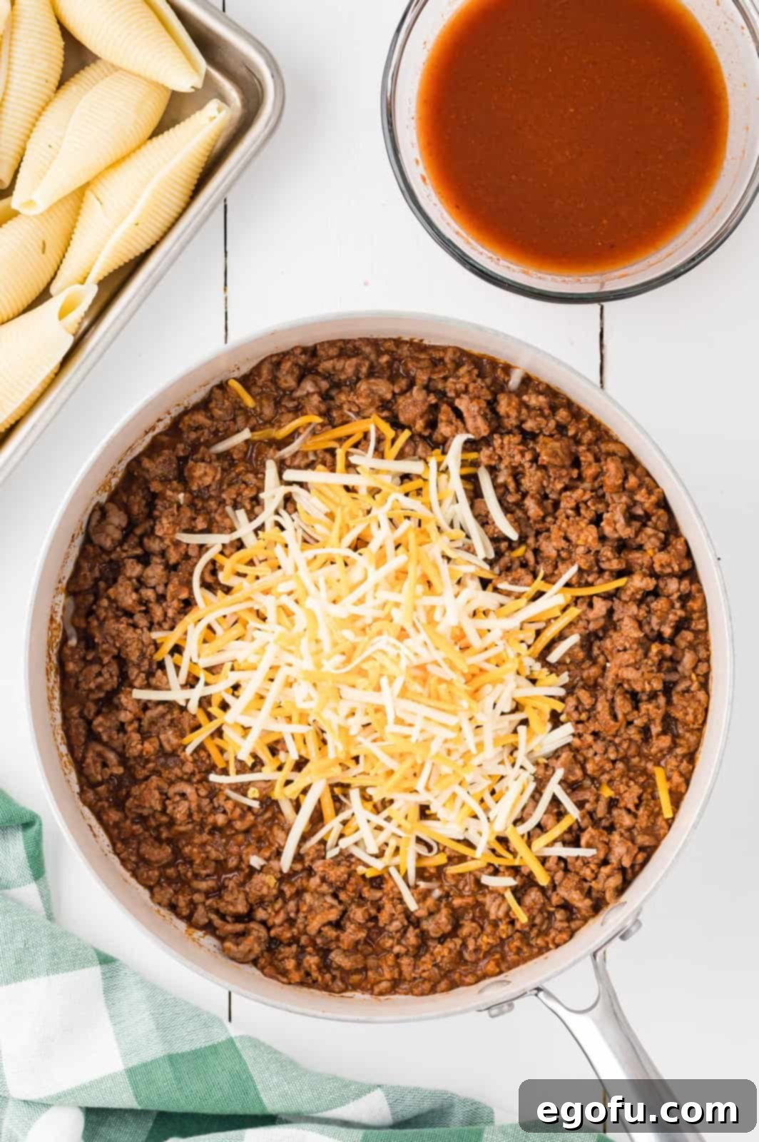 The taco meat mixture in a skillet, now with a generous sprinkling of shredded cheddar cheese, ready to be stirred in.