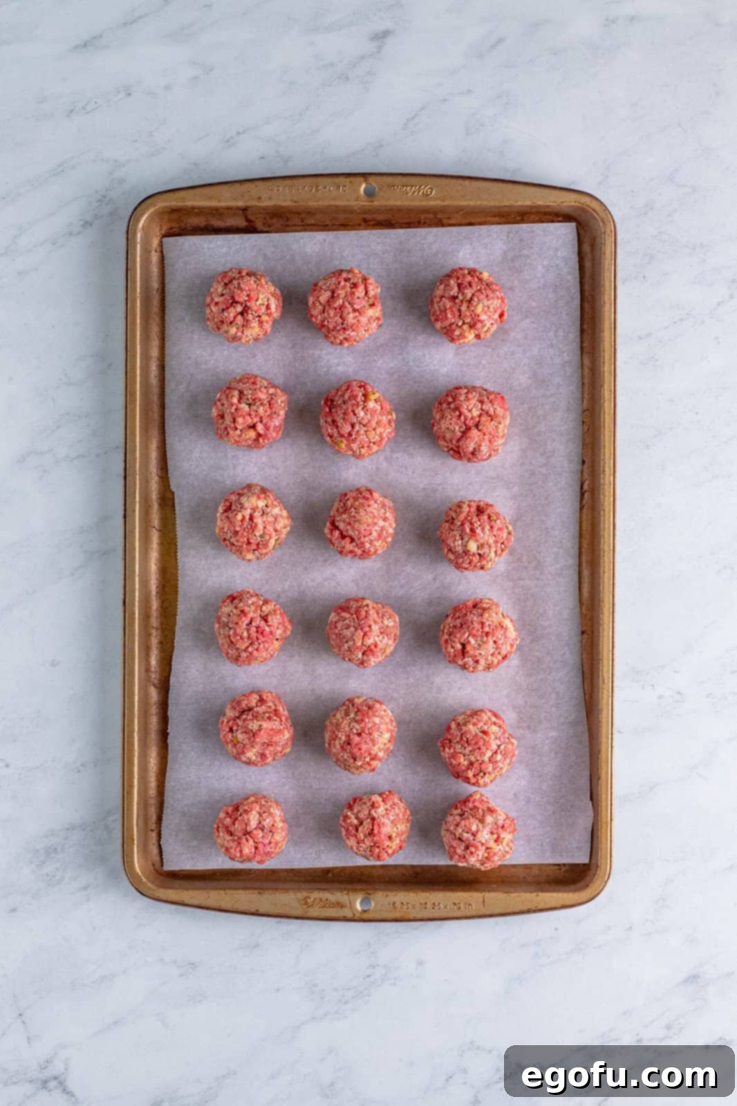 Small, uniformly-sized raw meatballs arranged neatly on a parchment-lined baking tray, ready for the oven.