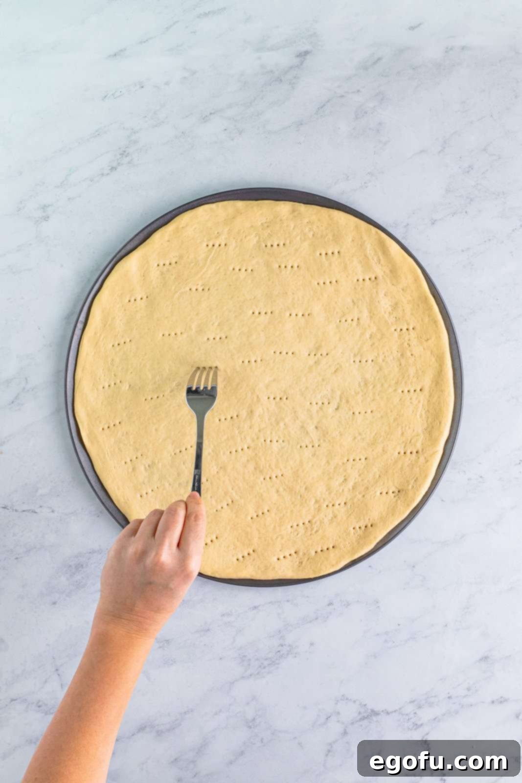A fork piercing holes across the surface of an unbaked pizza crust, a technique used for par-baking.