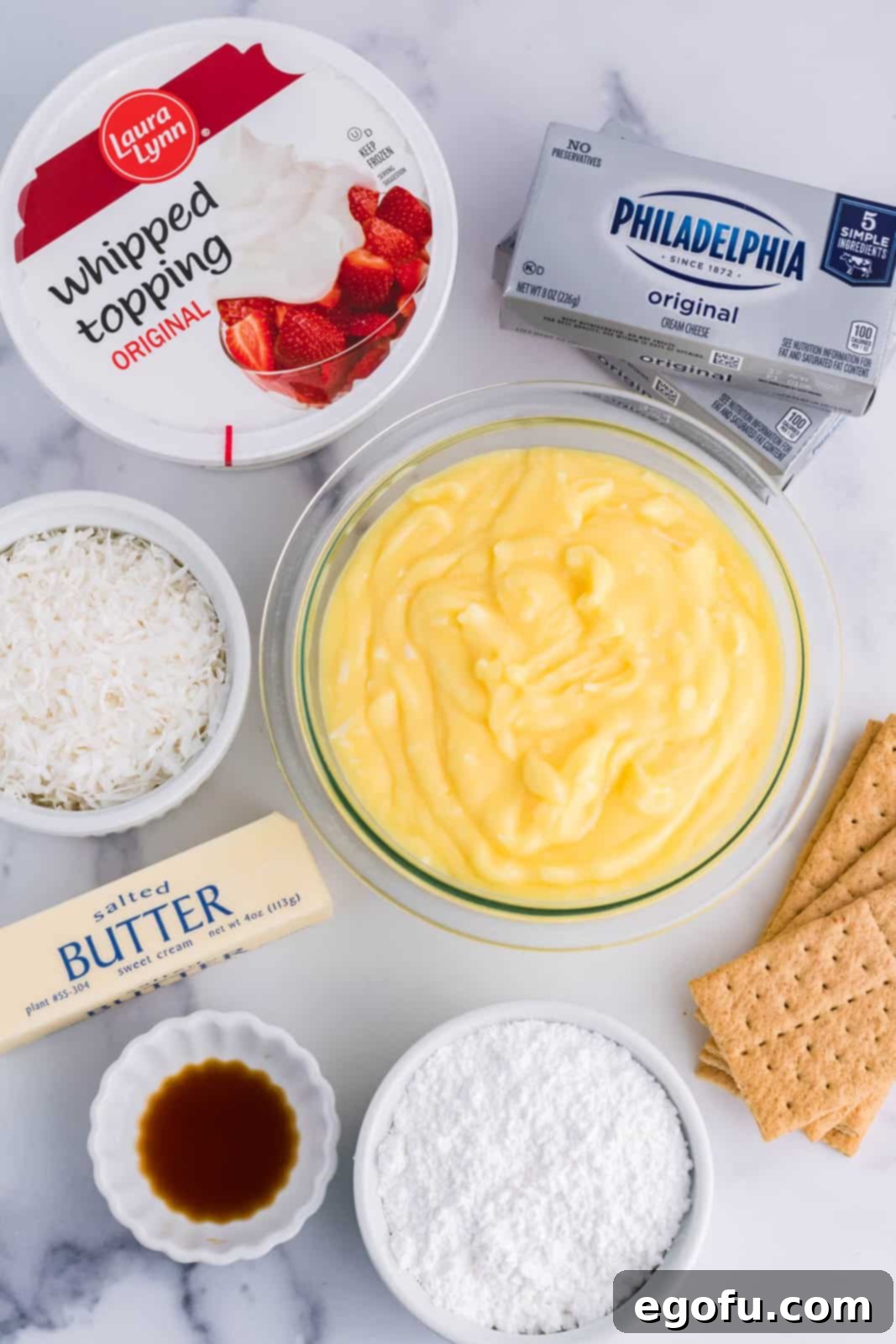 All the ingredients for Coconut Cream Lush laid out: coconut pudding, whipped topping, cream cheese, vanilla extract, butter, graham crackers, powdered sugar, milk, and coconut flakes.