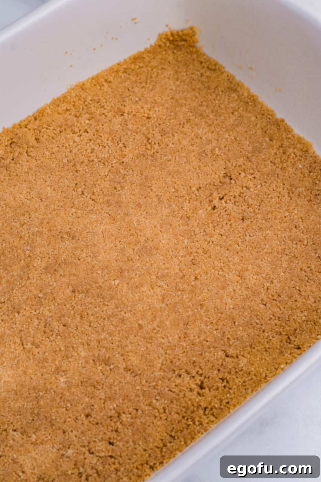 A golden brown graham cracker crust pressed evenly into the bottom of a clear glass baking pan.
