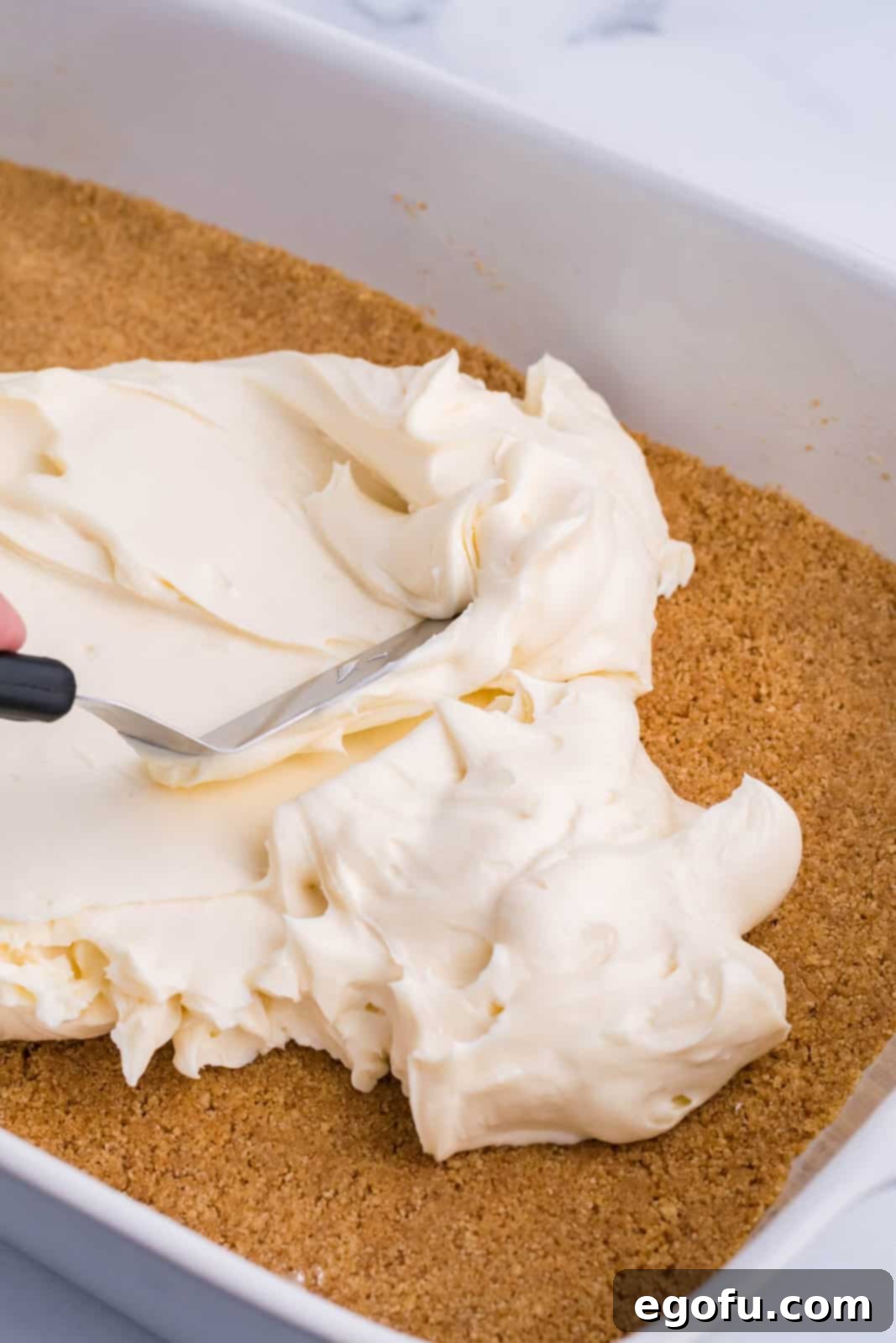A creamy, white cream cheese layer being spread smoothly over a graham cracker crust in a baking dish.