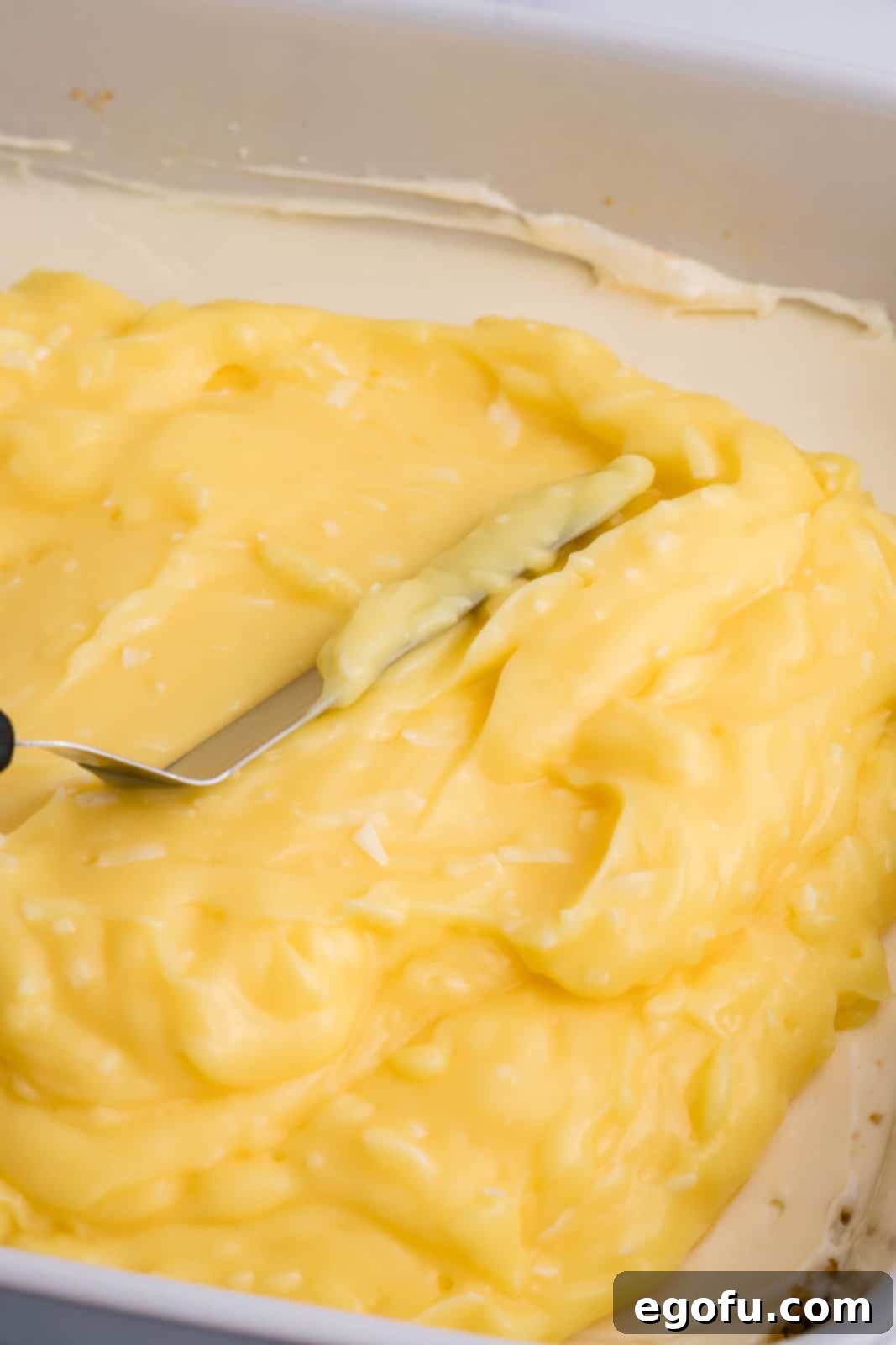 A thick, creamy layer of coconut pudding being spread evenly over the cream cheese layer of the dessert.