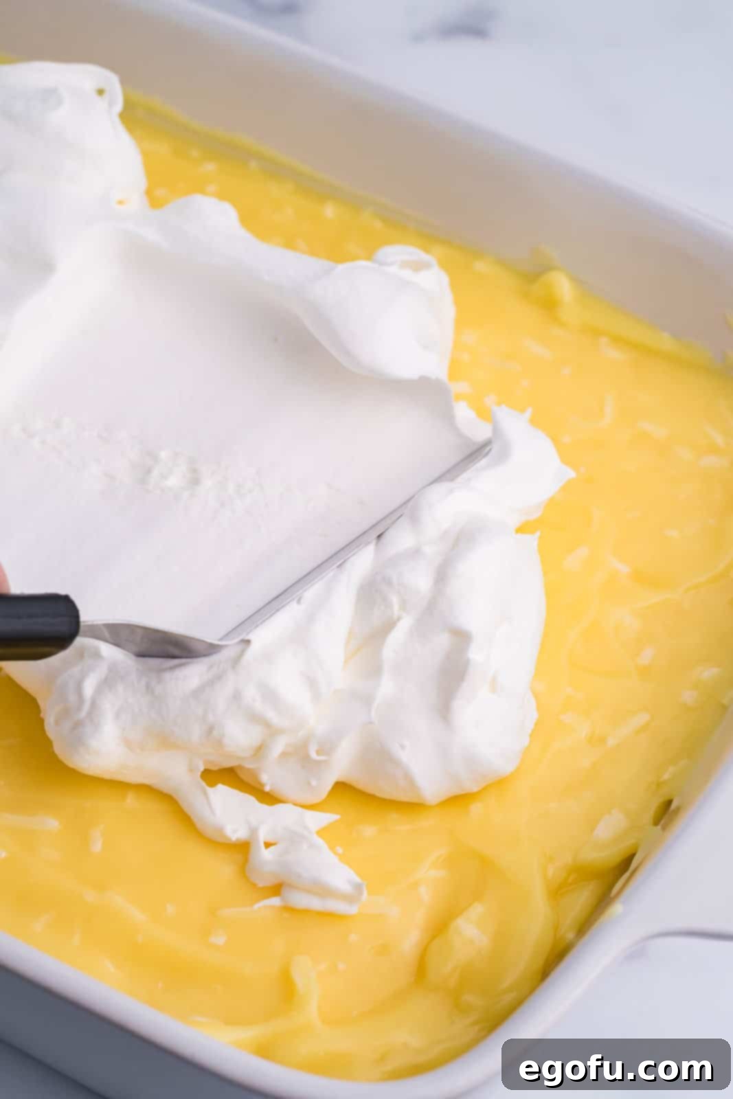 A fluffy white layer of whipped topping being spread smoothly over the coconut pudding layer in a baking dish.