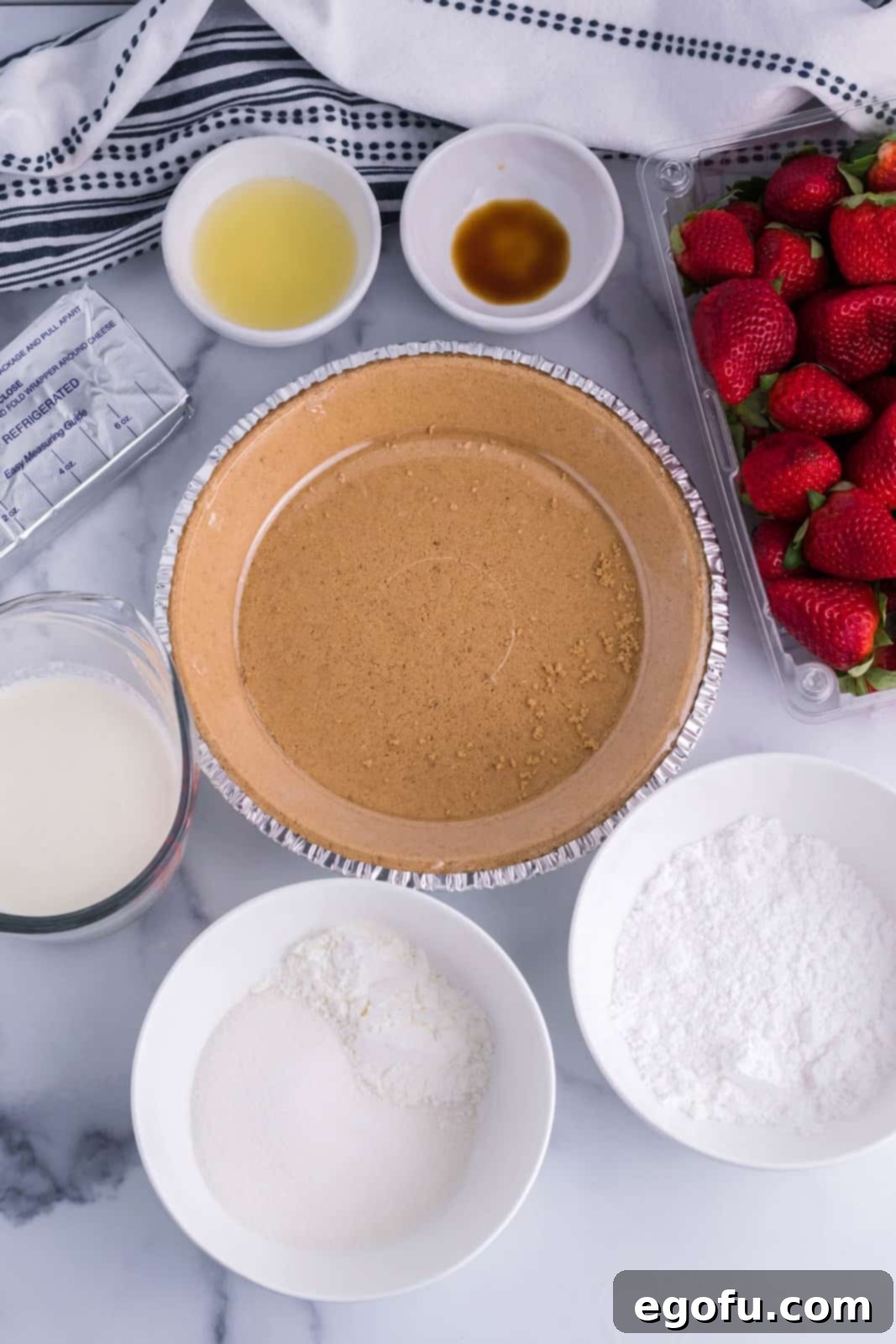 Individual ingredients for Strawberry Cream Cheese Pie laid out: graham cracker crust, sugar, cornstarch, lemon juice, cream cheese, powdered sugar, vanilla extract, and heavy cream.