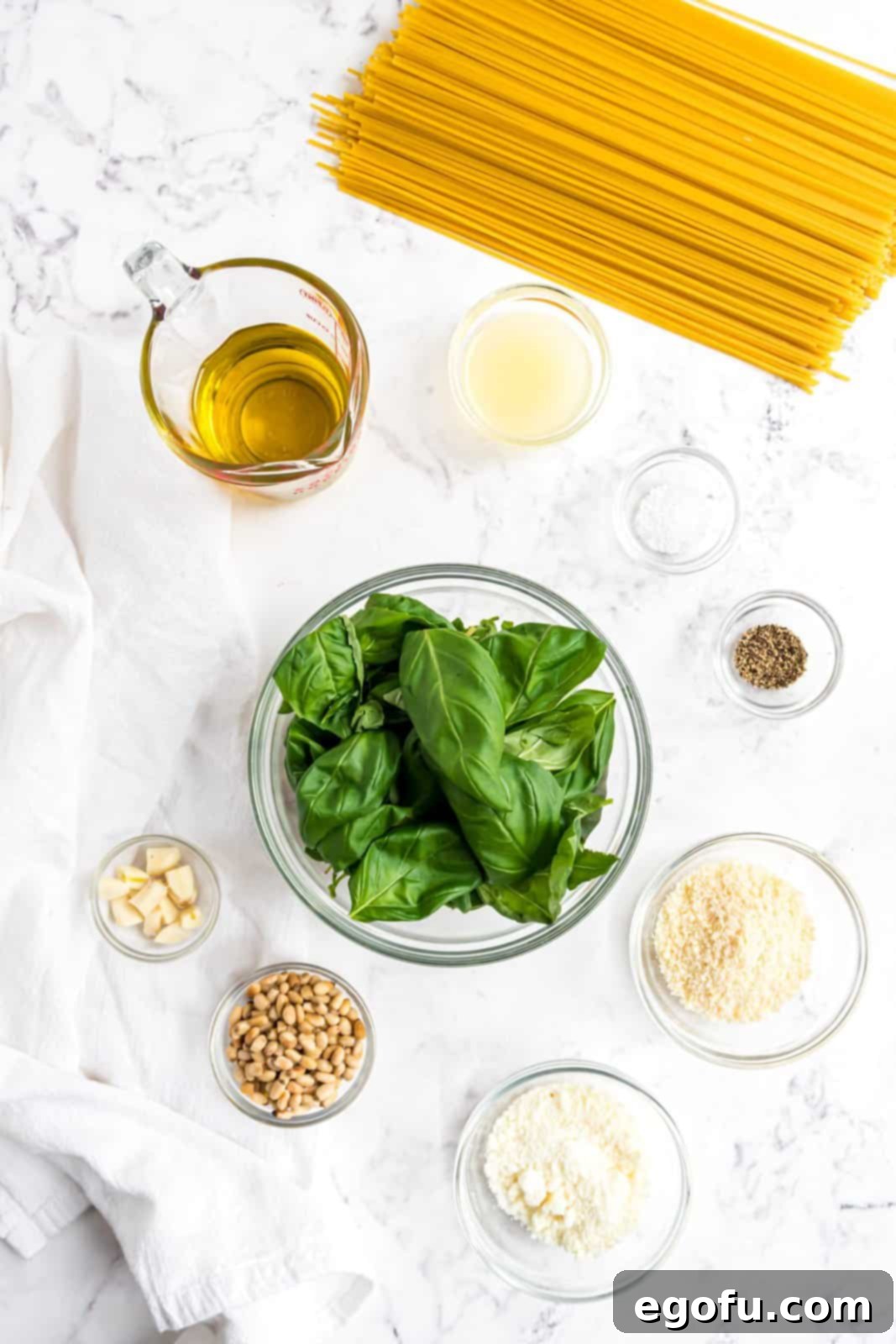 A beautiful arrangement of fresh ingredients for pesto pasta: basil leaves, kosher salt, spaghetti, garlic cloves, toasted pine nuts, olive oil, black pepper, lemon juice, pecorino romano cheese, and parmesan cheese, all ready for preparation.