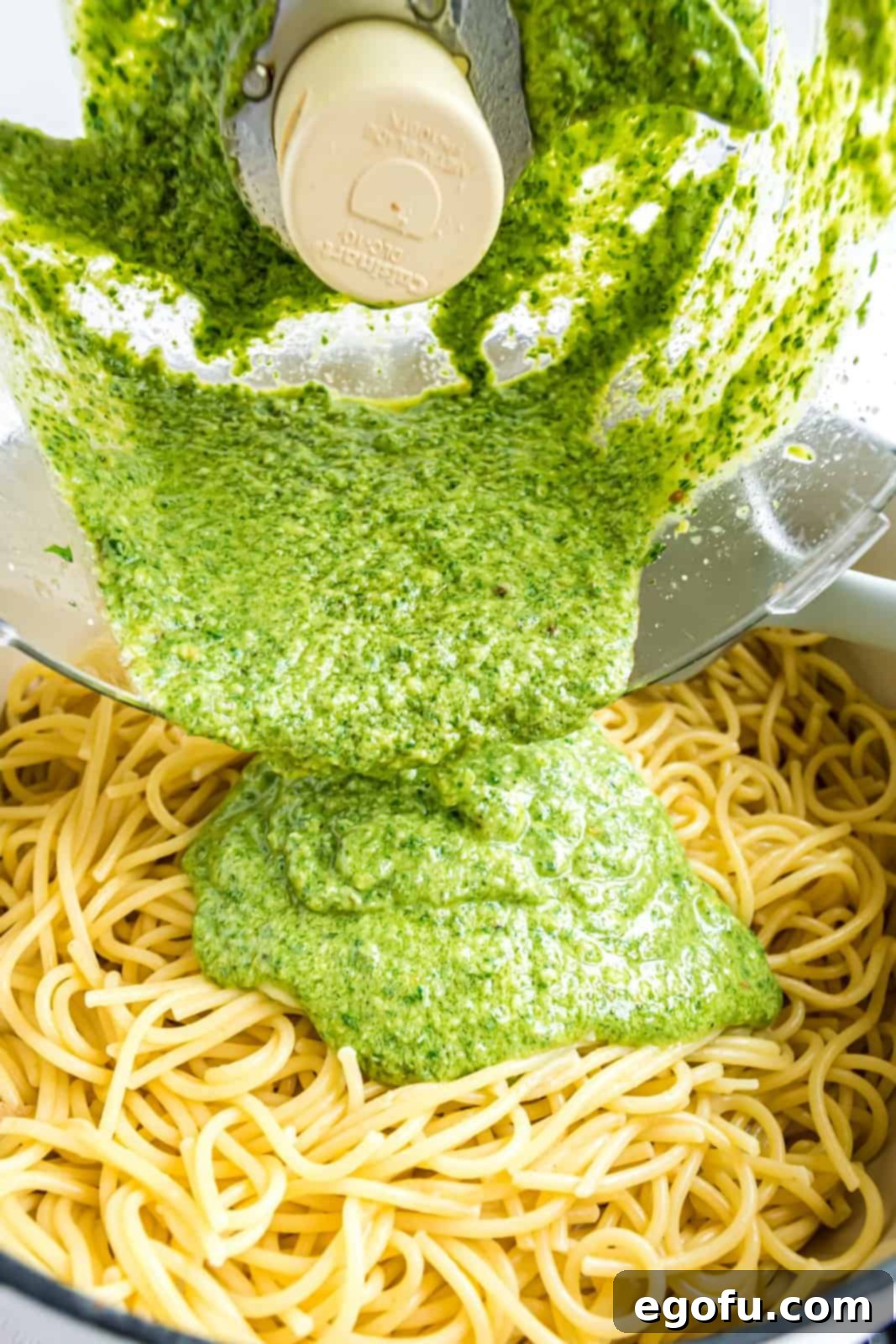 Vibrant fresh pesto sauce being generously poured over a bed of cooked spaghetti in a large bowl.