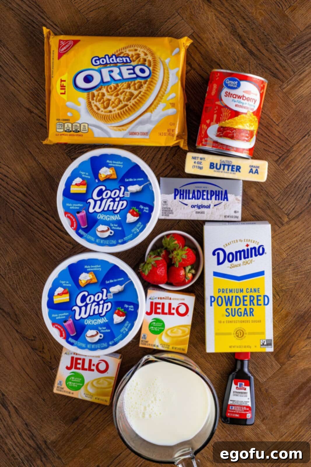 Layered Strawberry Bliss 4 A flat lay photograph showcasing all the ingredients for Strawberry Lasagna: Cool Whip, Golden Oreos, strawberry pie filling, vanilla pudding mix, cream cheese, butter, fresh strawberries, powdered sugar, and a glass of cold milk.