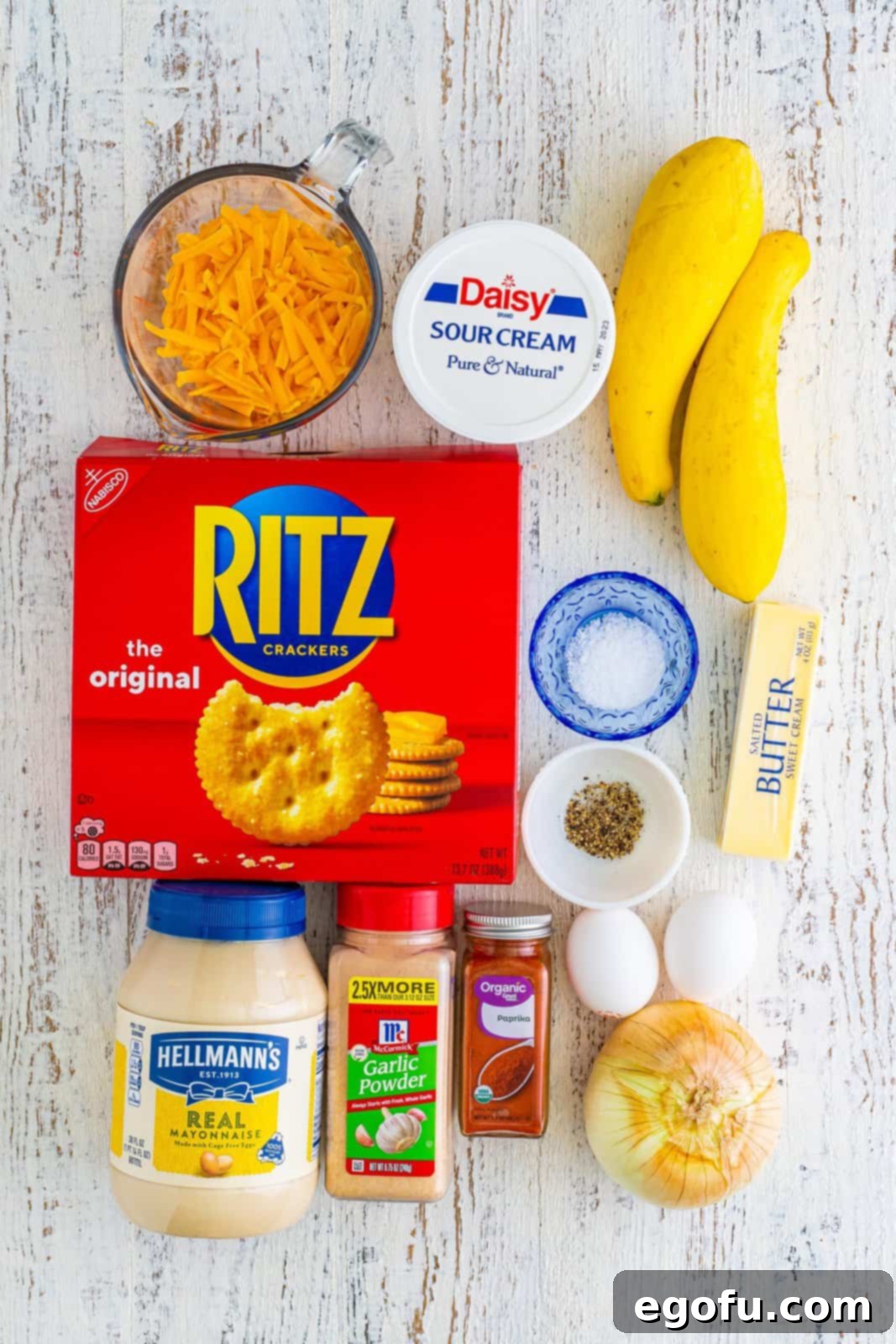 A vibrant display of all ingredients: buttery Ritz crackers, sliced yellow squash, creamy mayonnaise, sour cream, melted butter, sweet onion, kosher salt, black pepper, garlic powder, sweet paprika, large eggs, and shredded sharp cheddar cheese.