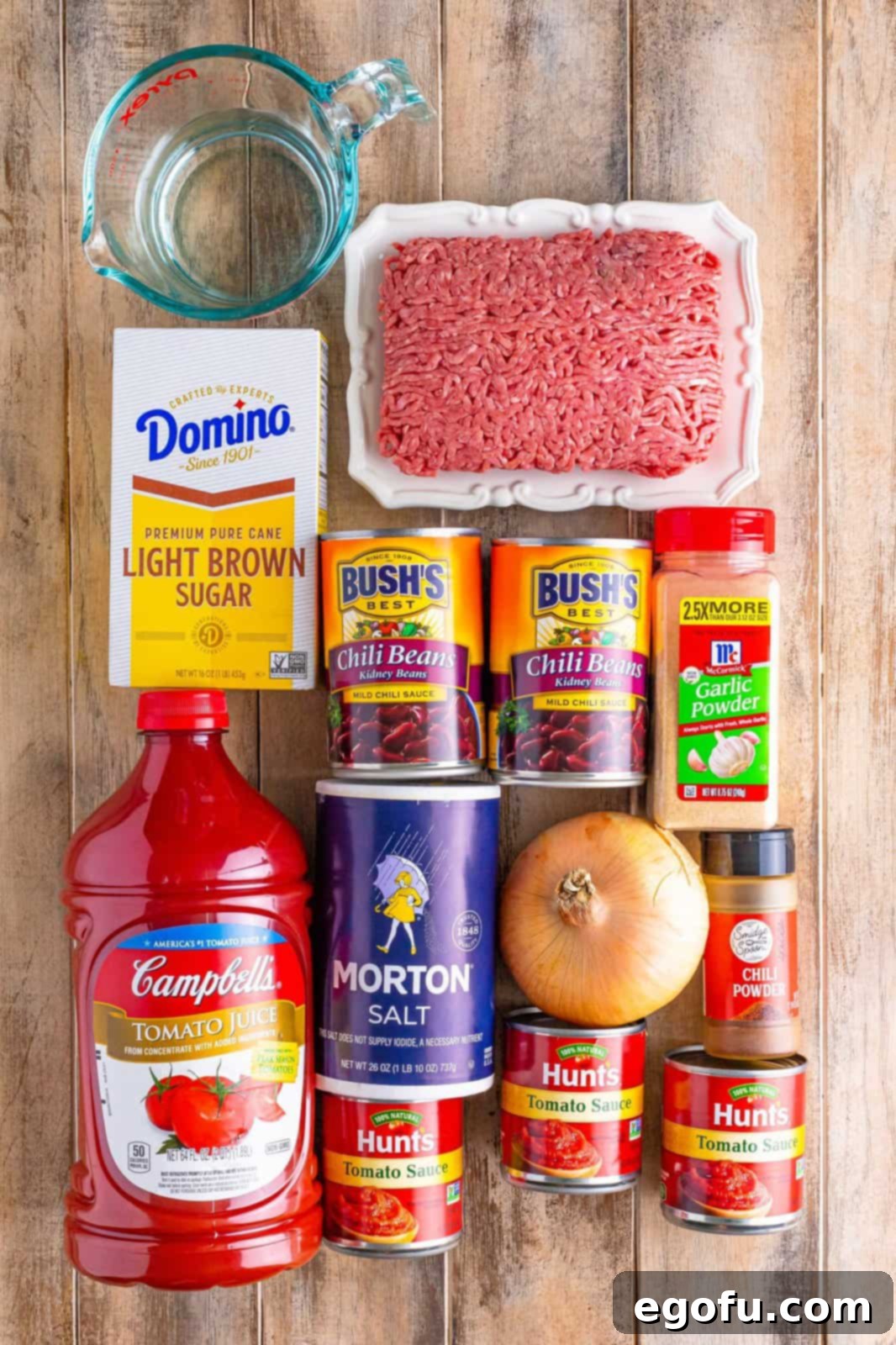 All the raw ingredients laid out neatly on a surface: ground beef, chopped onion, salt, light brown sugar, chili powder, garlic powder, cans of tomato sauce, tomato juice, water, and chili beans.