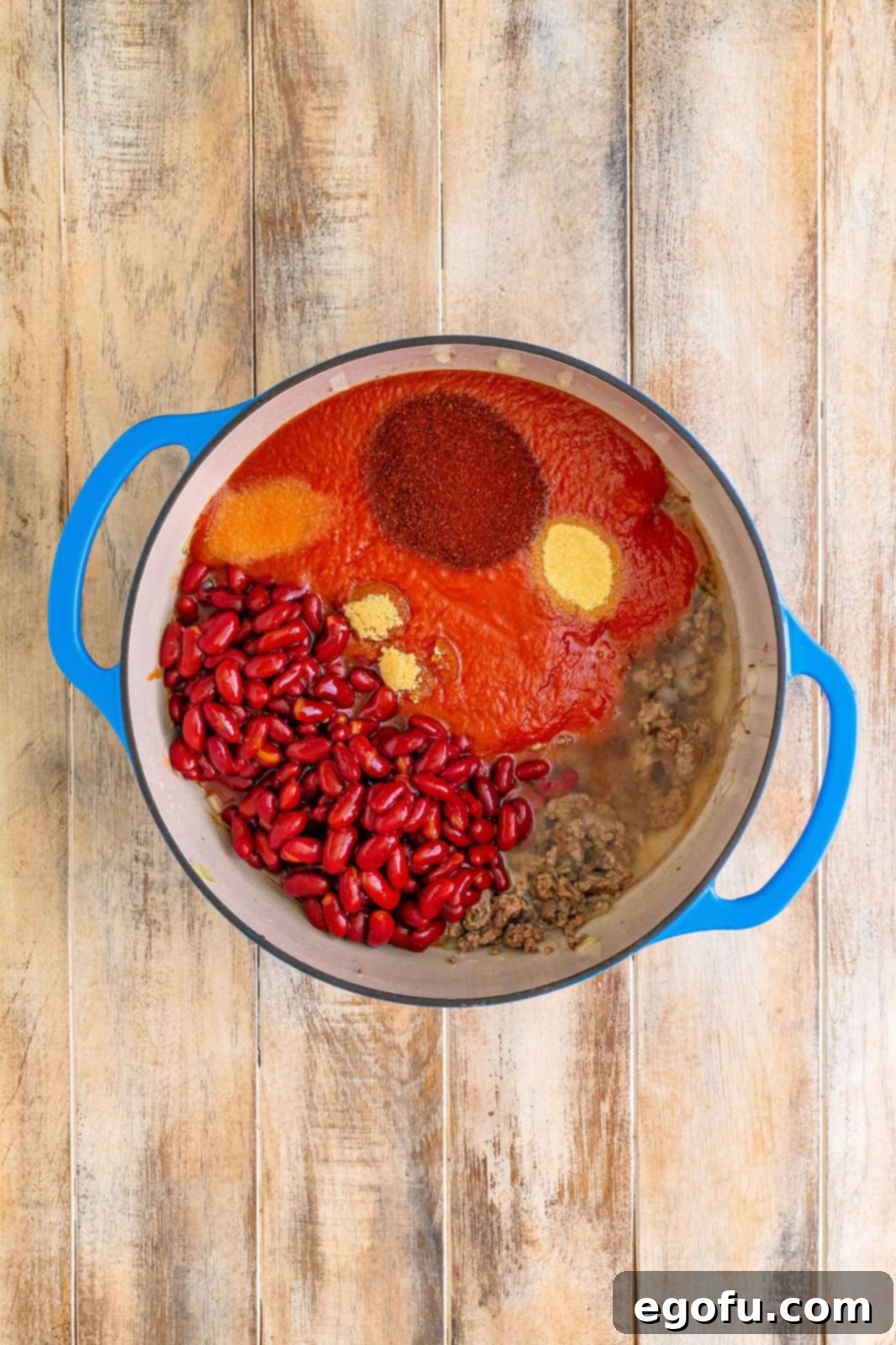 All chili ingredients, including tomato sauce, tomato juice, various spices, and chili beans, added to a Dutch oven with browned beef and onion, ready to be stirred.