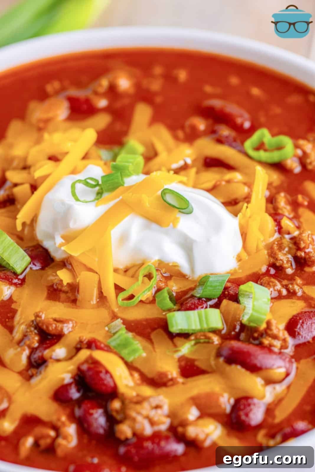 A close-up of a perfectly garnished bowl of rich cafeteria chili, topped with melted shredded cheese, a creamy dollop of sour cream, and vibrant chopped chives.