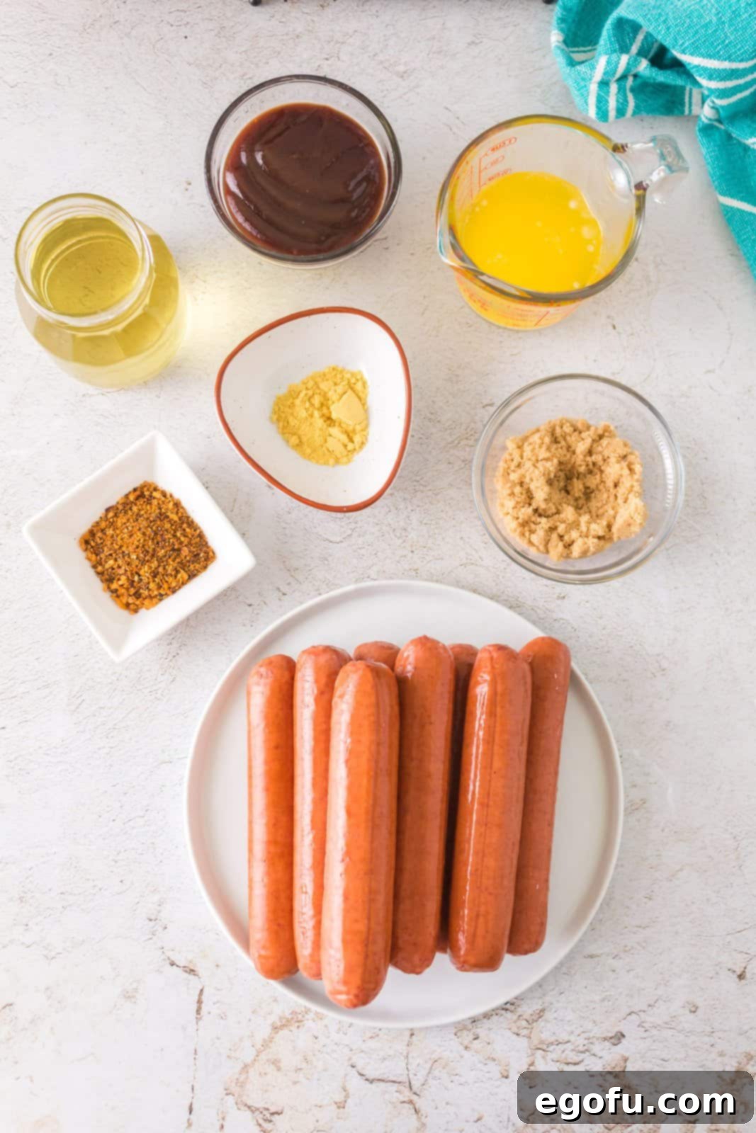 Hot dogs, dry mustard, olive oil, BBQ seasoning, BBQ sauce, butter, and brown sugar laid out on a table.
